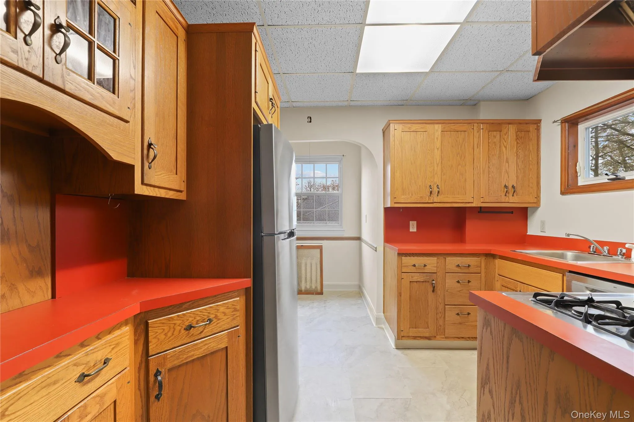 Kitchen featuring arched walkways, a drop ceiling, freestanding refrigerator, and brown cabinetry Kitchen featuring arched walkways, a drop ceiling, freestanding refrigerator, and brown cabinetry