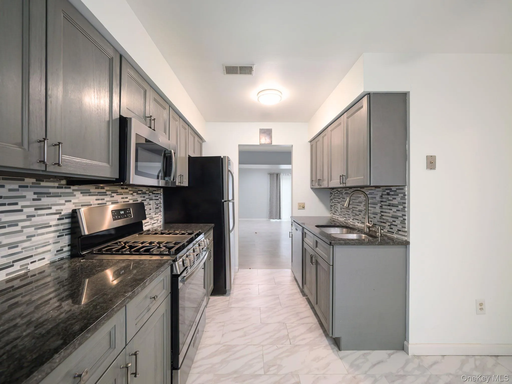 Kitchen featuring appliances with stainless steel finishes, dark stone counters, light marble finish flooring, gray cabinetry, and decorative backsplash Kitchen featuring appliances with stainless steel finishes, dark stone counters, light marble finish flooring, gray cabinetry, and decorative backsplash