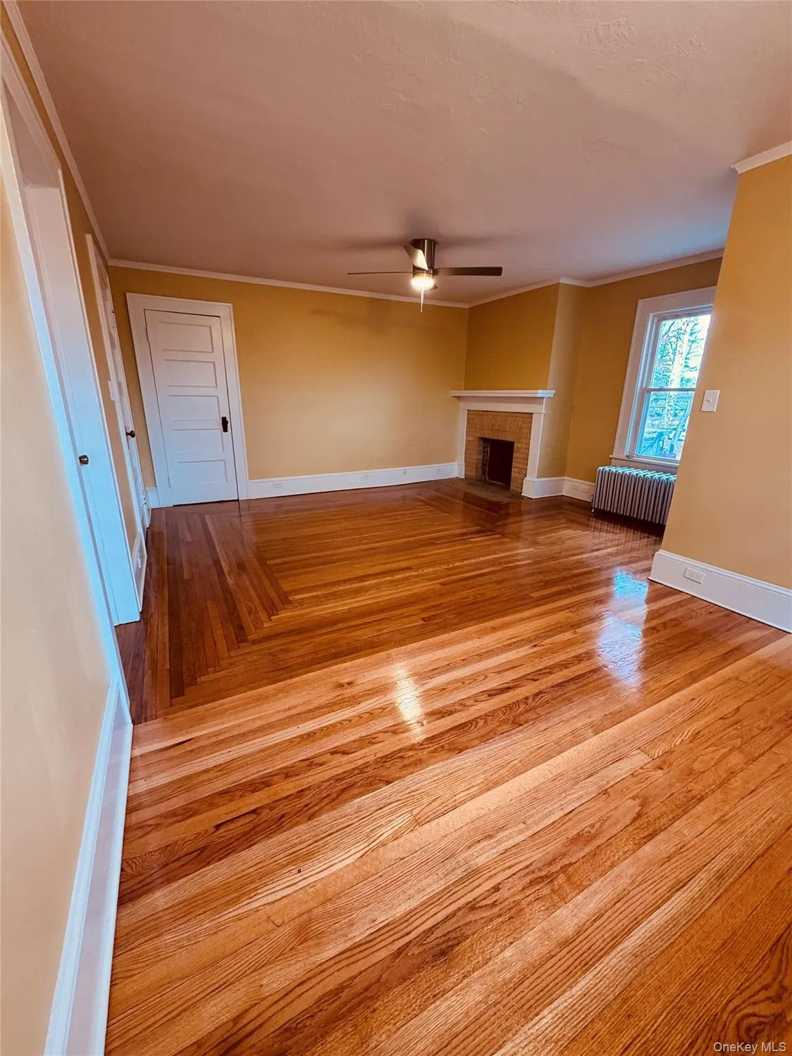 Unfurnished living room featuring crown molding, a brick fireplace, light wood-style floors, radiator, and ceiling fan Unfurnished living room featuring crown molding, a brick fireplace, light wood-style floors, radiator, and ceiling fan