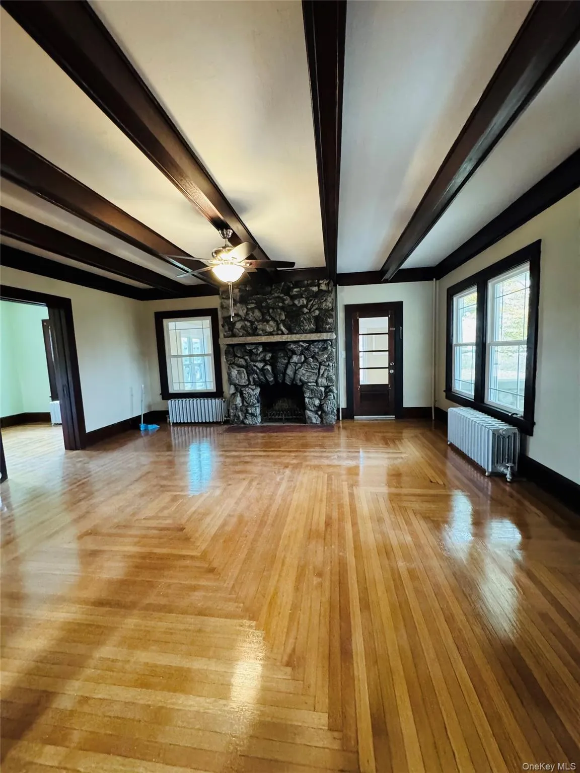 Unfurnished living room with beam ceiling, a stone fireplace, radiator, ceiling fan, and parquet floors Unfurnished living room with beam ceiling, a stone fireplace, radiator, ceiling fan, and parquet floors