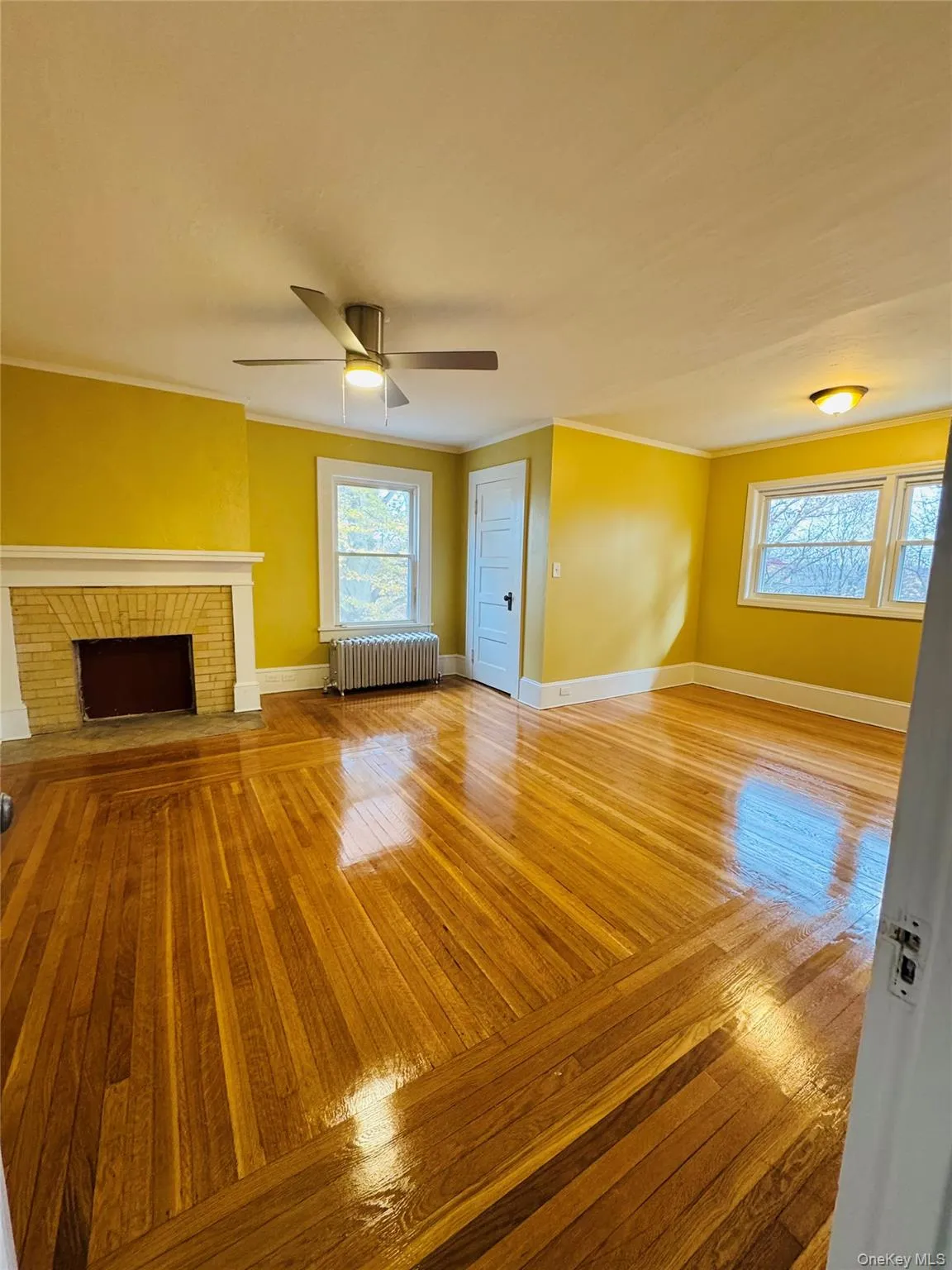 Unfurnished living room with light wood-style flooring, radiator heating unit, a brick fireplace, a ceiling fan, and crown molding Unfurnished living room with light wood-style flooring, radiator heating unit, a brick fireplace, a ceiling fan, and crown molding