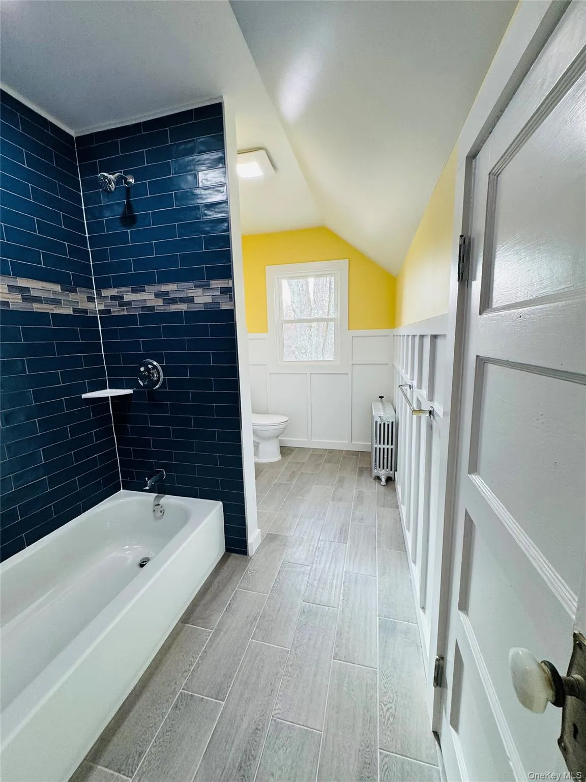 Bathroom with wainscoting, tub / shower combination, vaulted ceiling, radiator, and wood finish floors Bathroom with wainscoting, tub / shower combination, vaulted ceiling, radiator, and wood finish floors