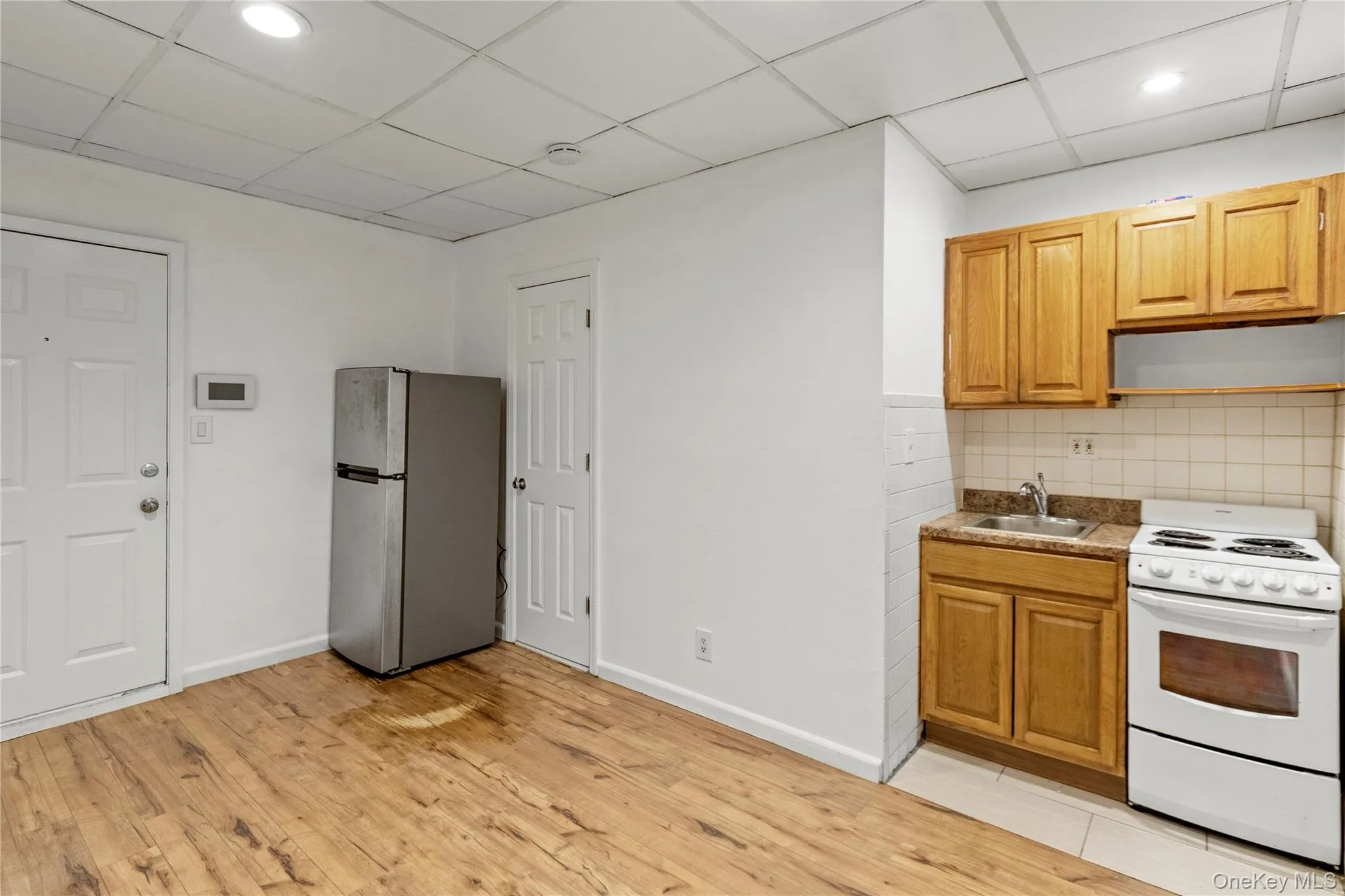 Kitchen featuring white range with electric stovetop, a drop ceiling, freestanding refrigerator, extractor fan, and brown cabinets Kitchen featuring white range with electric stovetop, a drop ceiling, freestanding refrigerator, extractor fan, and brown cabinets