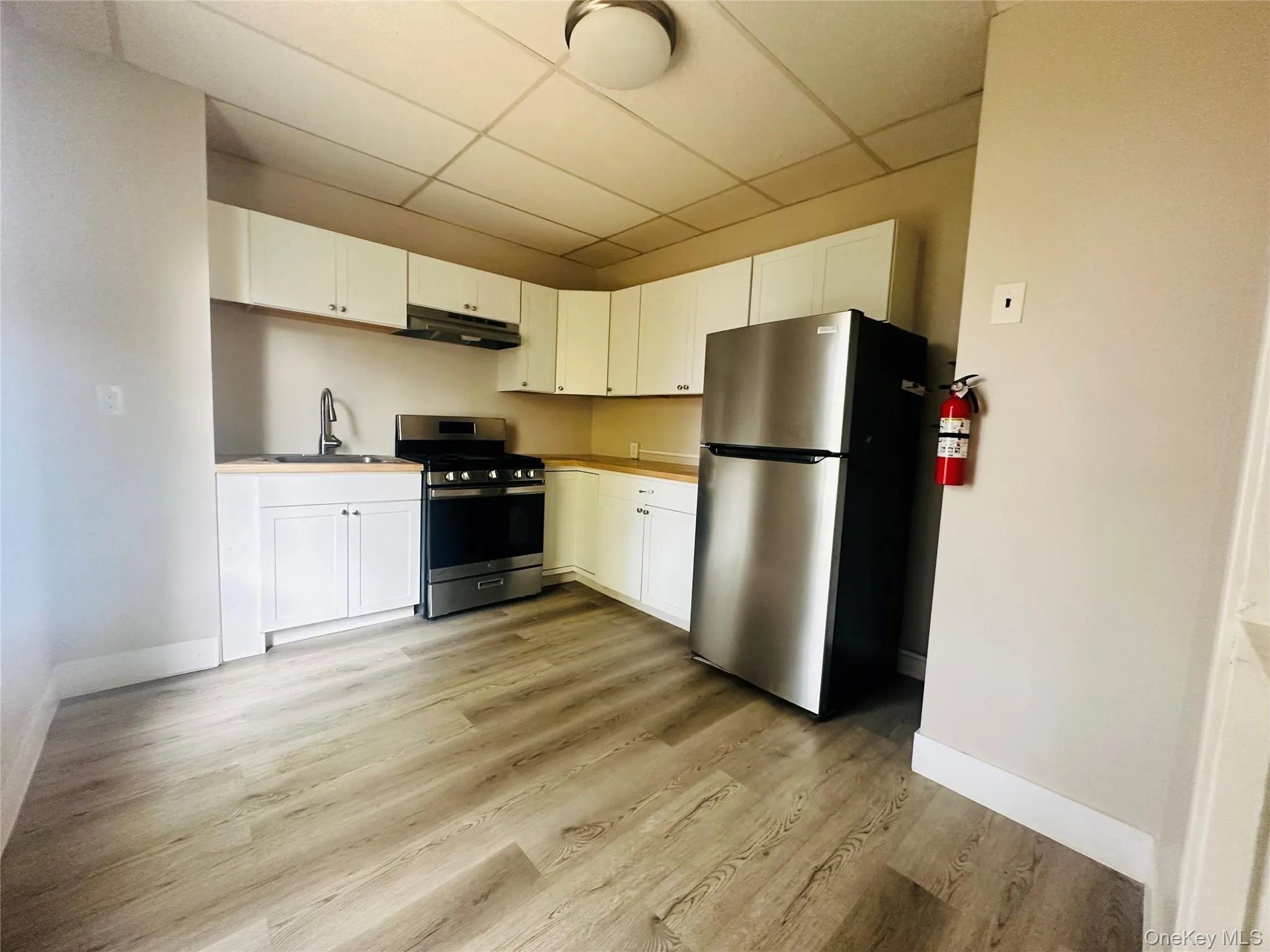 Kitchen featuring appliances with stainless steel finishes, a paneled ceiling, white cabinets, light countertops, and light wood finished floors Kitchen featuring appliances with stainless steel finishes, a paneled ceiling, white cabinets, light countertops, and light wood finished floors