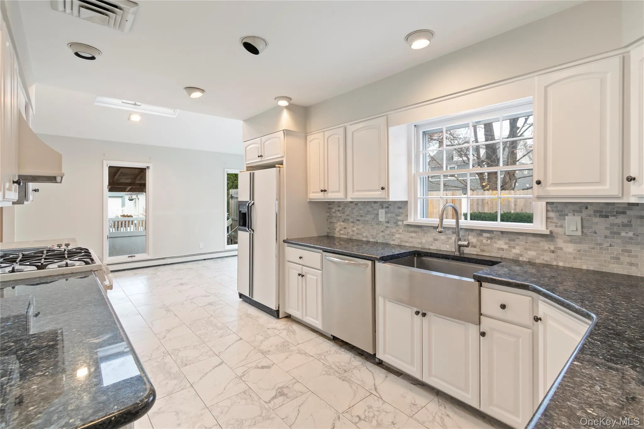 Kitchen featuring light marble finish flooring, dark stone countertops, white refrigerator with ice dispenser, tasteful backsplash, and stainless steel dishwasher Kitchen featuring light marble finish flooring, dark stone countertops, white refrigerator with ice dispenser, tasteful backsplash, and stainless steel dishwasher