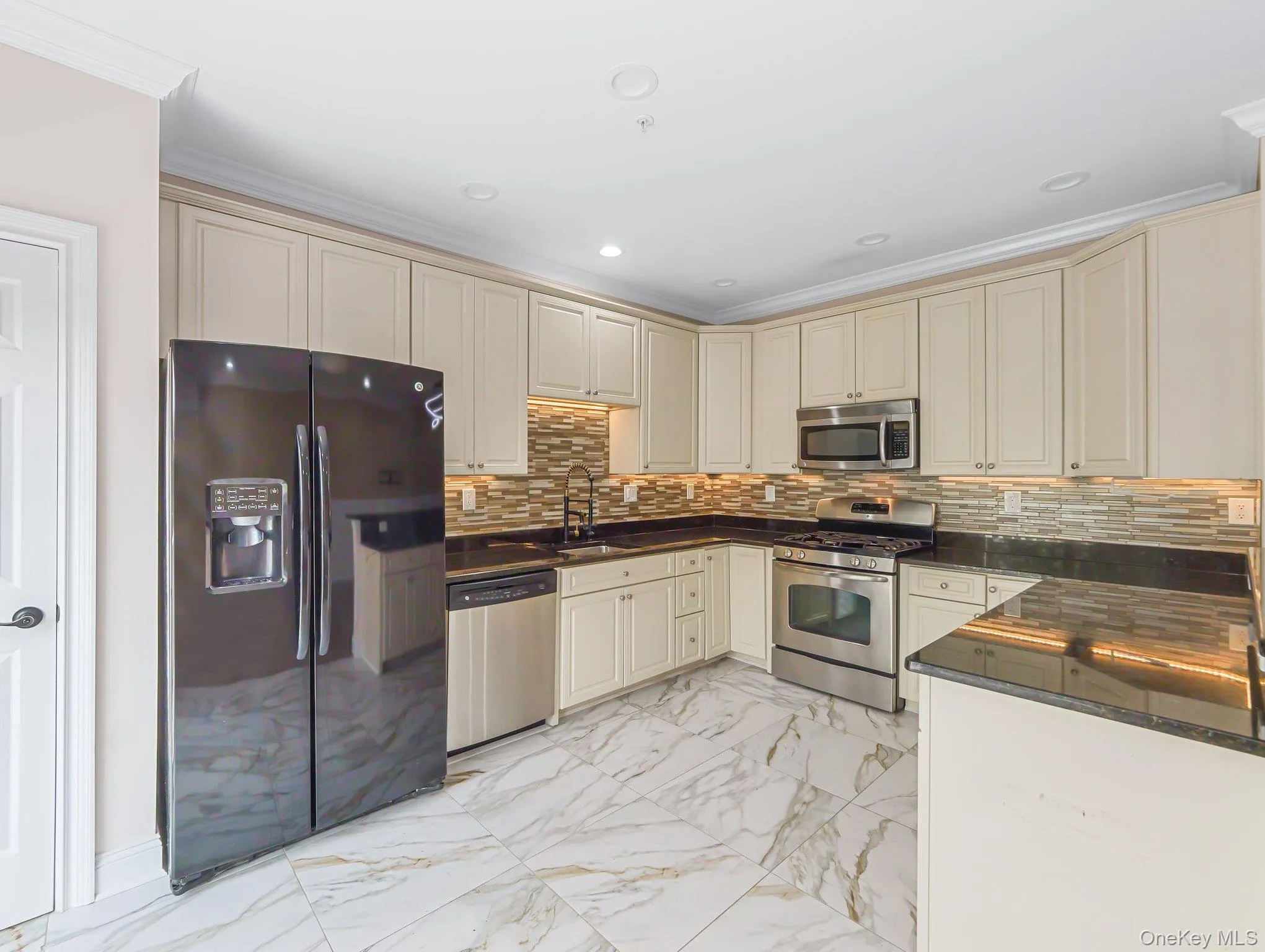 Kitchen featuring cream cabinets, stainless steel appliances, light marble finish floors, dark stone counters, and crown molding Kitchen featuring cream cabinets, stainless steel appliances, light marble finish floors, dark stone counters, and crown molding