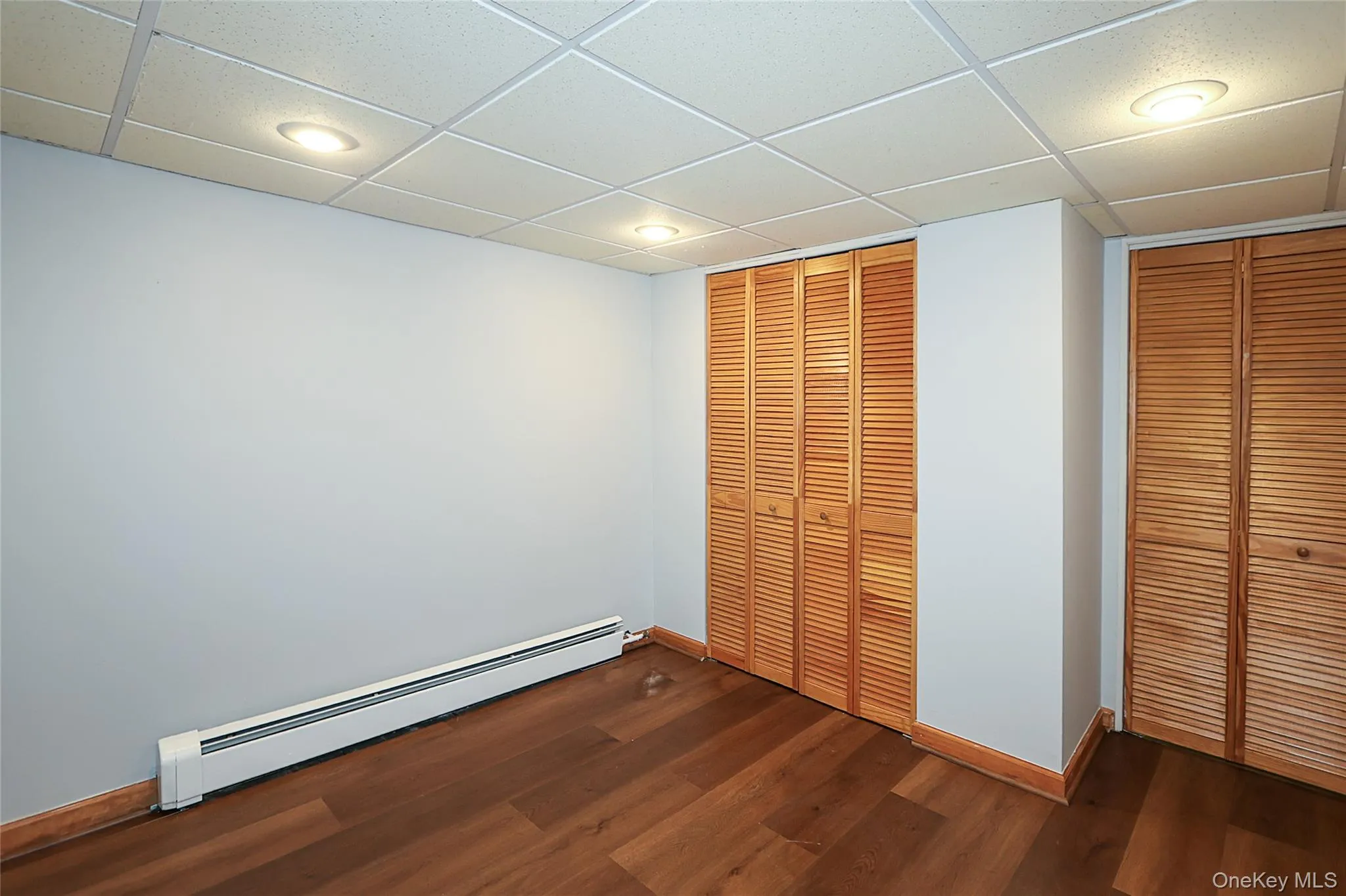 Unfurnished bedroom featuring a paneled ceiling, baseboard heating, dark wood-style floors, two closets, and recessed lighting Unfurnished bedroom featuring a paneled ceiling, baseboard heating, dark wood-style floors, two closets, and recessed lighting