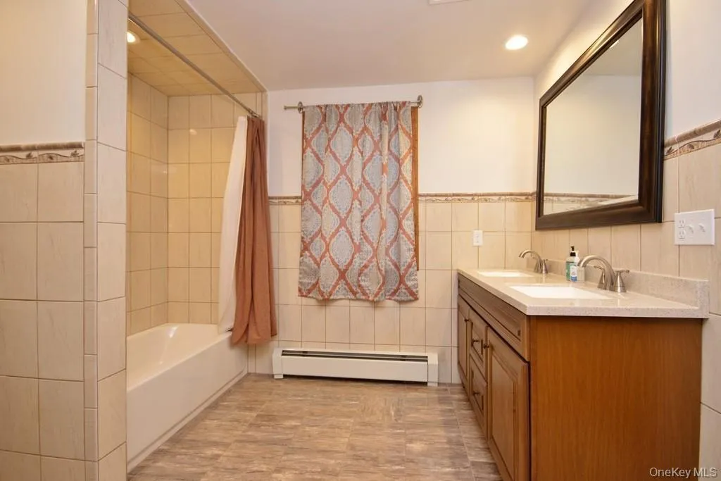 Bathroom featuring tile walls, shower / tub combo, baseboard heating, double vanity, and wainscoting Bathroom featuring tile walls, shower / tub combo, baseboard heating, double vanity, and wainscoting