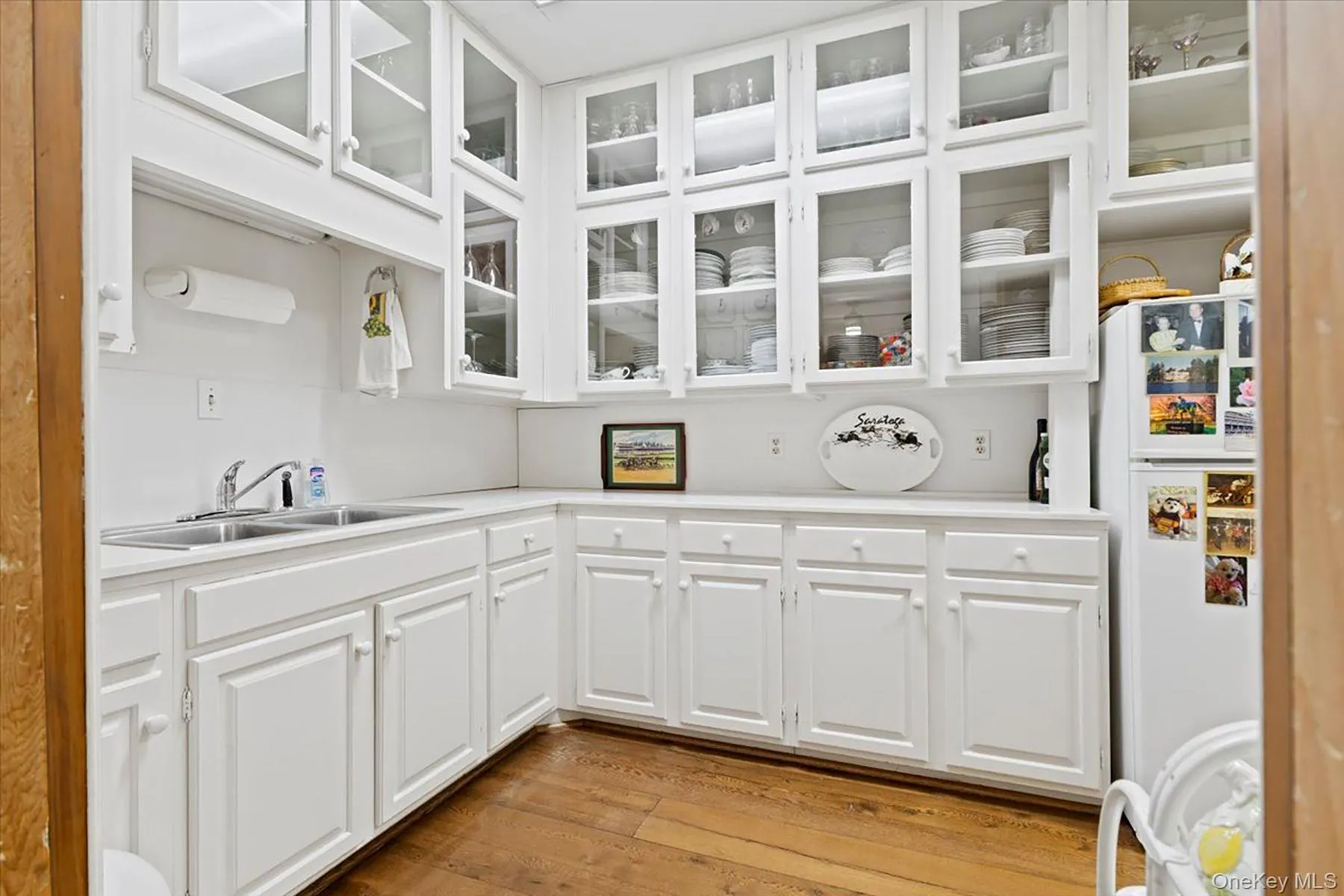 Kitchen featuring white cabinets, light countertops, light wood-type flooring, freestanding refrigerator, and glass insert cabinets Kitchen featuring white cabinets, light countertops, light wood-type flooring, freestanding refrigerator, and glass insert cabinets