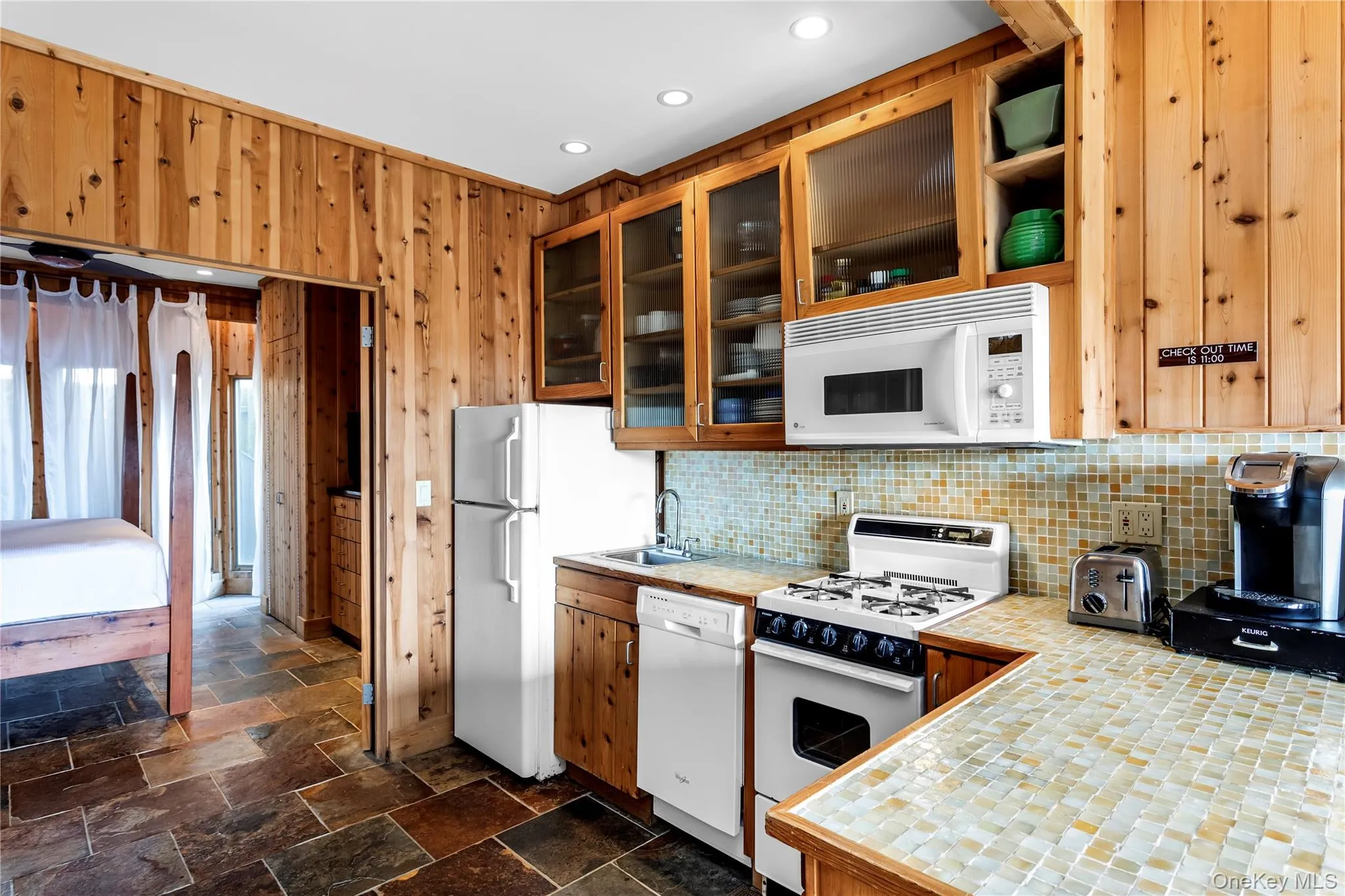 Kitchen featuring white appliances, tasteful backsplash, glass insert cabinets, stone tile floors, and wooden walls Kitchen featuring white appliances, tasteful backsplash, glass insert cabinets, stone tile floors, and wooden walls