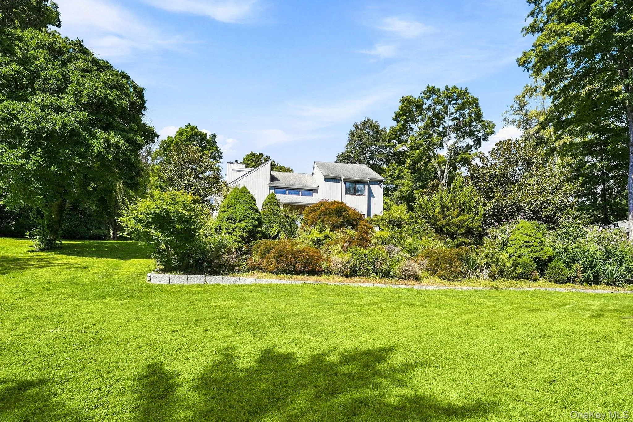 Expansive lawn area with a stone-edged planting bed, surrounded by mature trees and shrubs, featuring a multi-level architectural design Expansive lawn area with a stone-edged planting bed, surrounded by mature trees and shrubs, featuring a multi-level architectural design