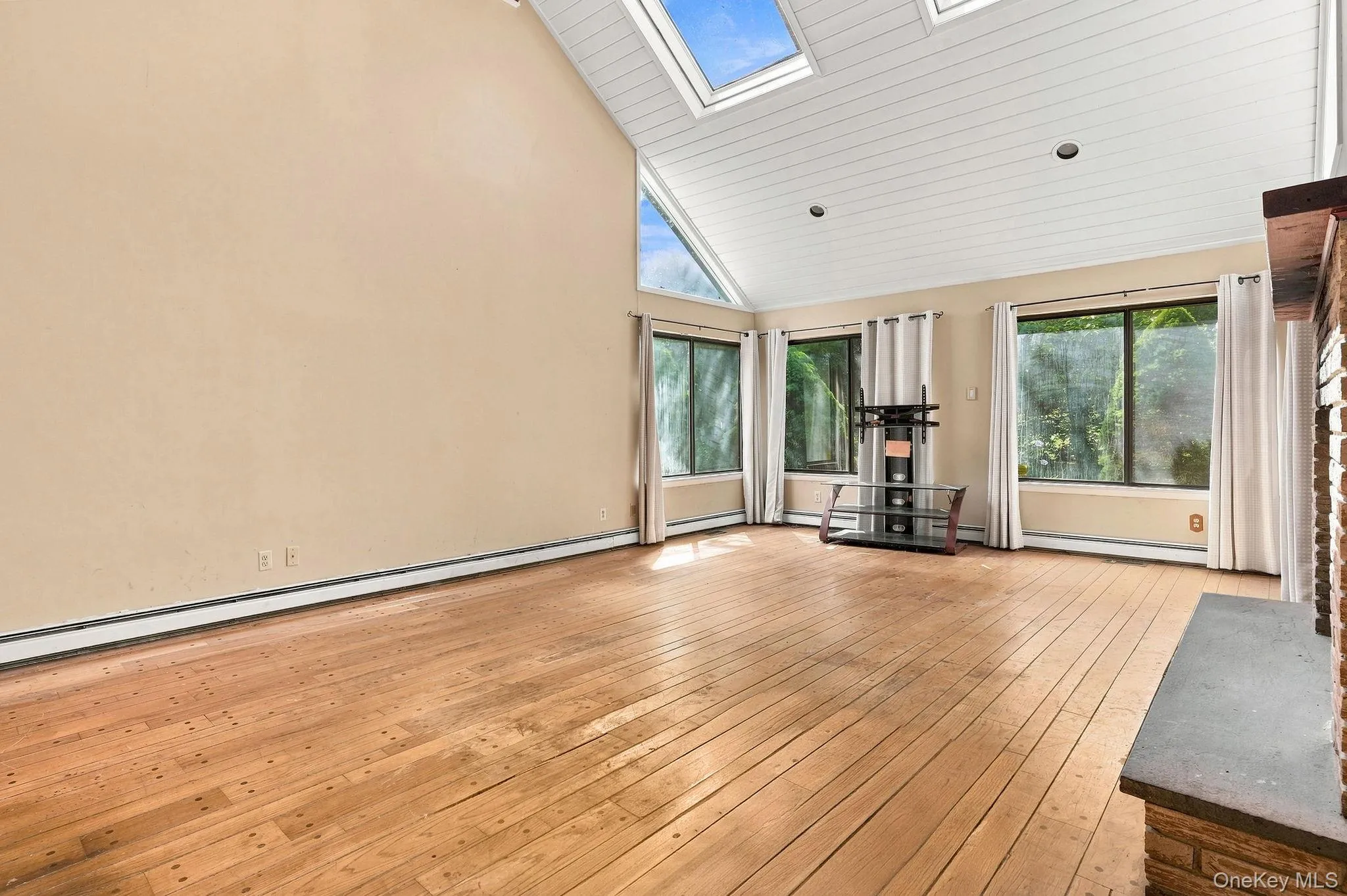 Vaulted ceiling with skylights, wood-finish flooring, multiple large windows, and a brick-finish fireplace with a stone hearth Vaulted ceiling with skylights, wood-finish flooring, multiple large windows, and a brick-finish fireplace with a stone hearth
