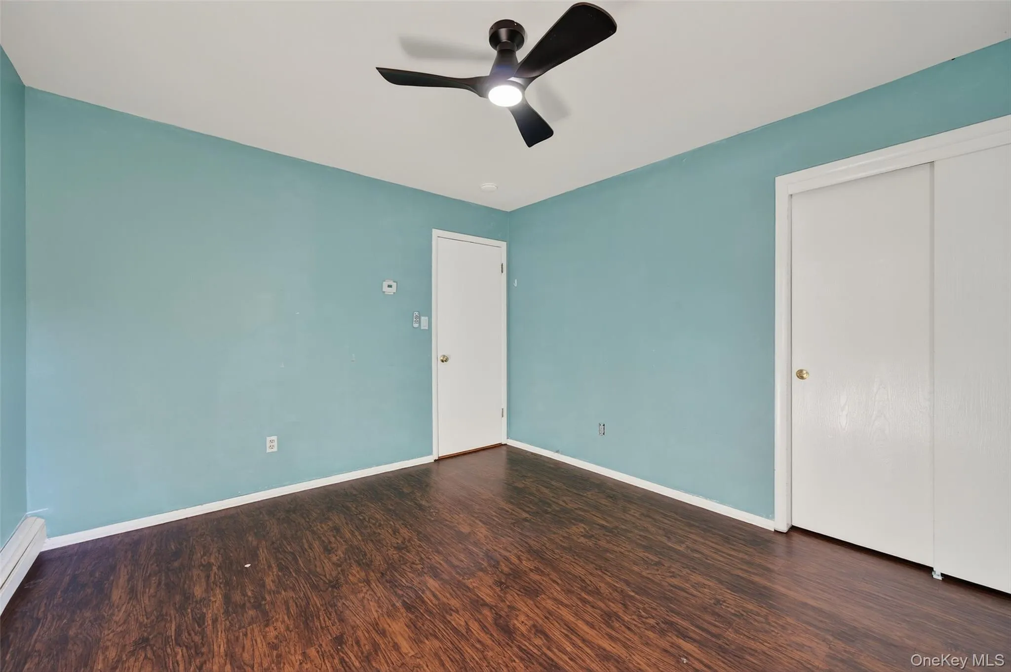 Unfurnished bedroom with dark wood-style flooring, a ceiling fan, a baseboard heating unit, and a closet Unfurnished bedroom with dark wood-style flooring, a ceiling fan, a baseboard heating unit, and a closet