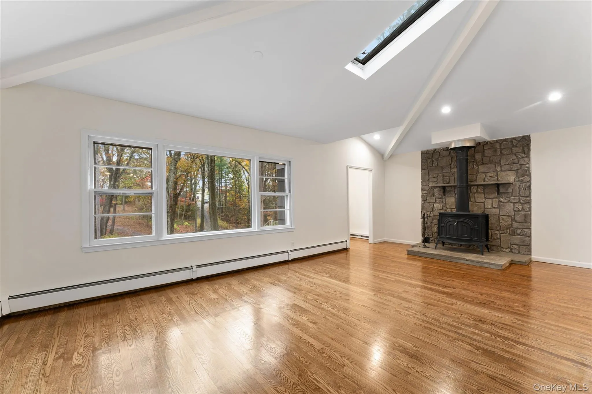 54 De Liso: Unfurnished living room with a wood stove, a baseboard heating unit, light wood-type flooring, beamed ceiling, and high vaulted ceiling 54 De Liso: Unfurnished living room with a wood stove, a baseboard heating unit, light wood-type flooring, beamed ceiling, and high vaulted ceiling