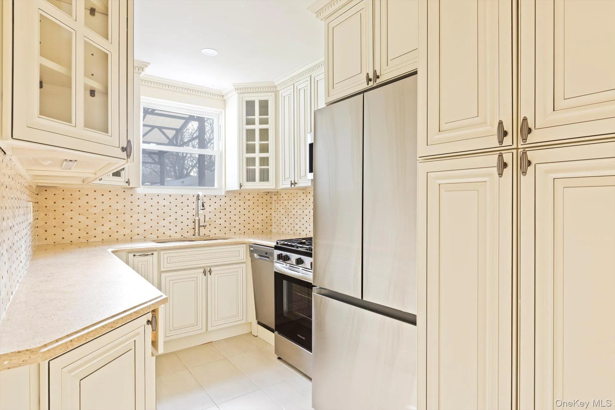 Kitchen featuring appliances with stainless steel finishes, light countertops, cream cabinets, glass insert cabinets, and tasteful backsplash Kitchen featuring appliances with stainless steel finishes, light countertops, cream cabinets, glass insert cabinets, and tasteful backsplash