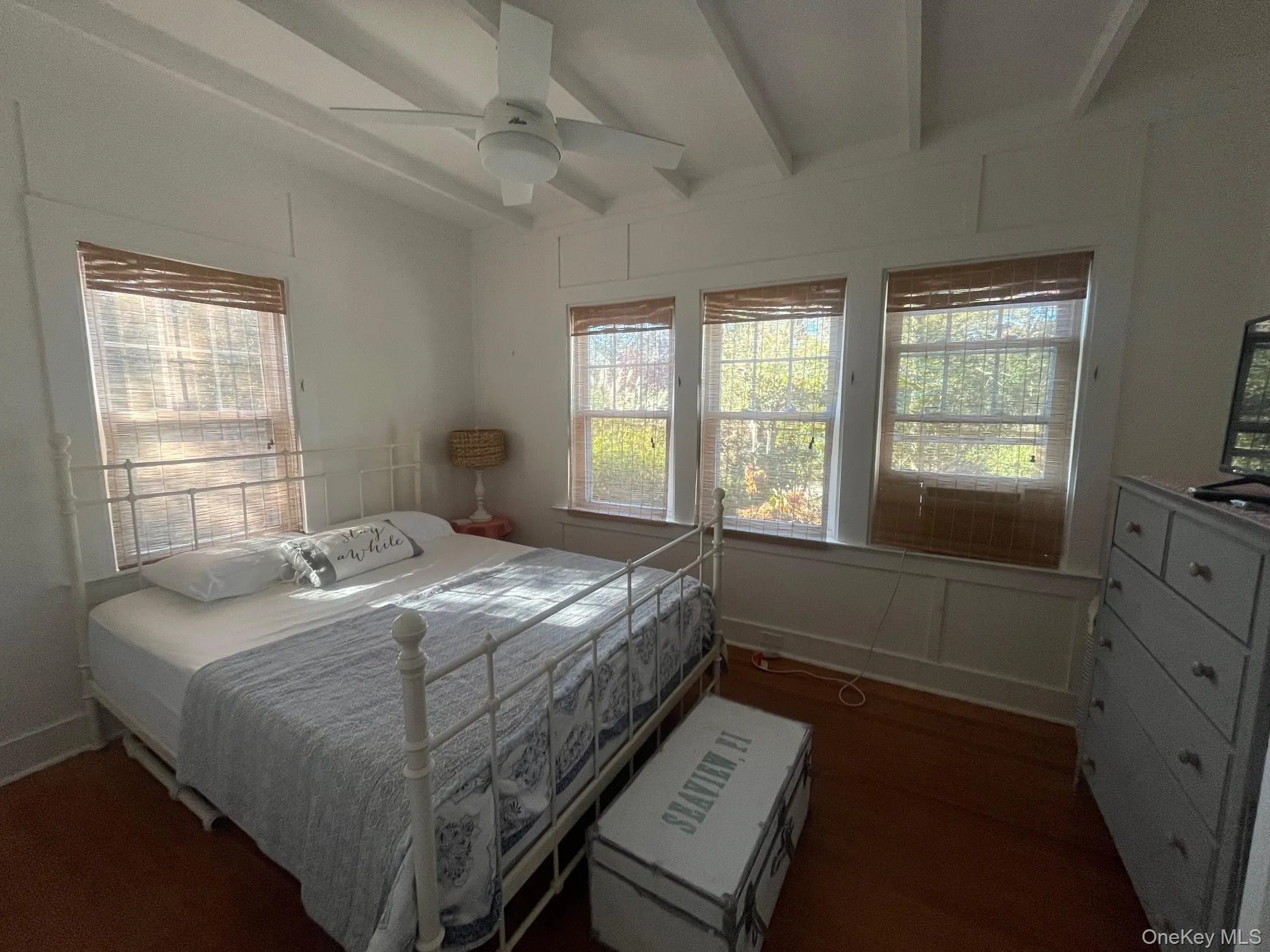 Bedroom with beam ceiling, multiple windows, dark wood-style flooring, ceiling fan, and a decorative wall Bedroom with beam ceiling, multiple windows, dark wood-style flooring, ceiling fan, and a decorative wall