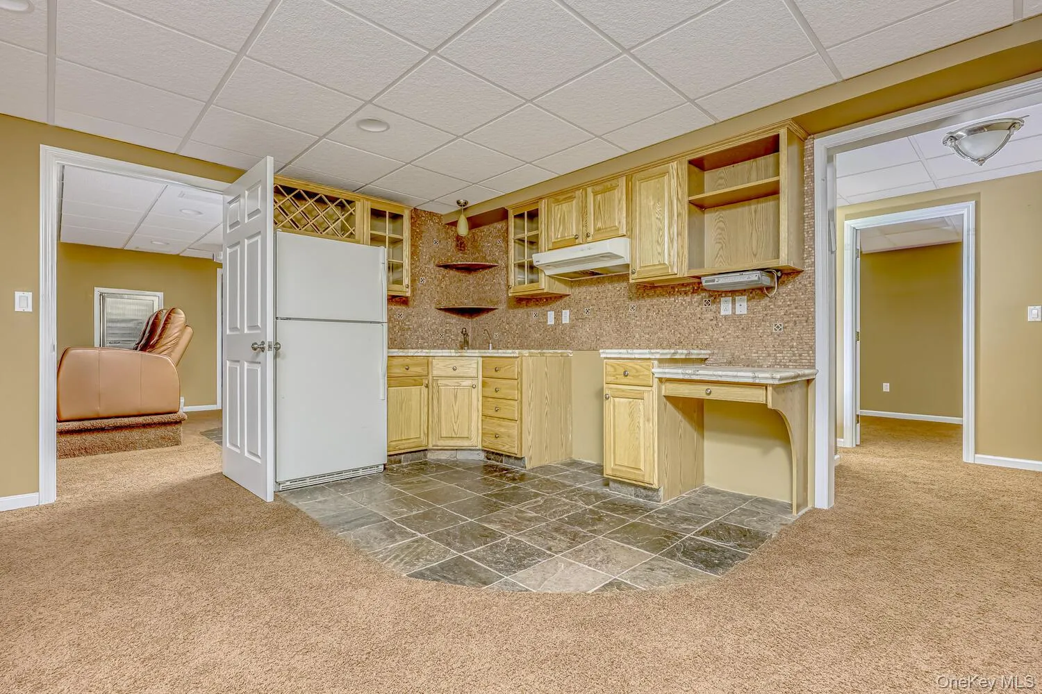 Kitchen featuring a drop ceiling, light carpet, freestanding refrigerator, light brown cabinetry, and light countertops Kitchen featuring a drop ceiling, light carpet, freestanding refrigerator, light brown cabinetry, and light countertops