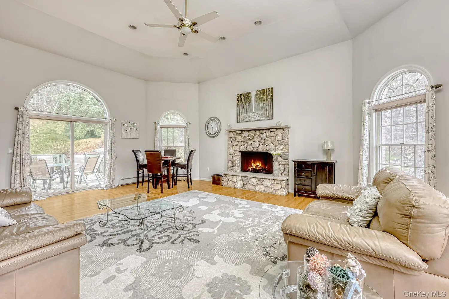 Living room with a stone fireplace, light wood-type flooring, ceiling fan, plenty of natural light, and a high ceiling Living room with a stone fireplace, light wood-type flooring, ceiling fan, plenty of natural light, and a high ceiling