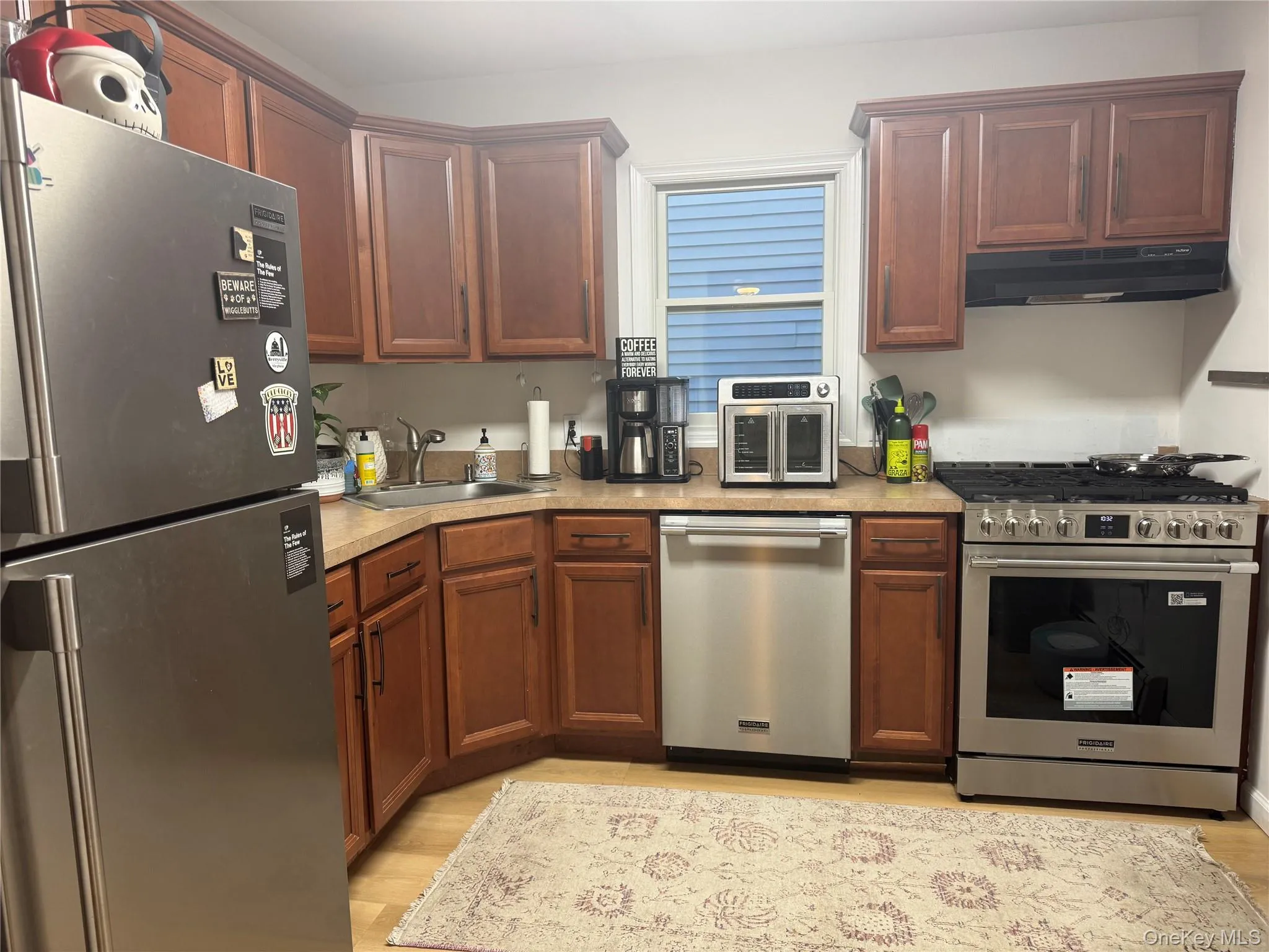 Kitchen featuring stainless steel appliances, light wood-style floors, light countertops, under cabinet range hood, and brown cabinetry Kitchen featuring stainless steel appliances, light wood-style floors, light countertops, under cabinet range hood, and brown cabinetry