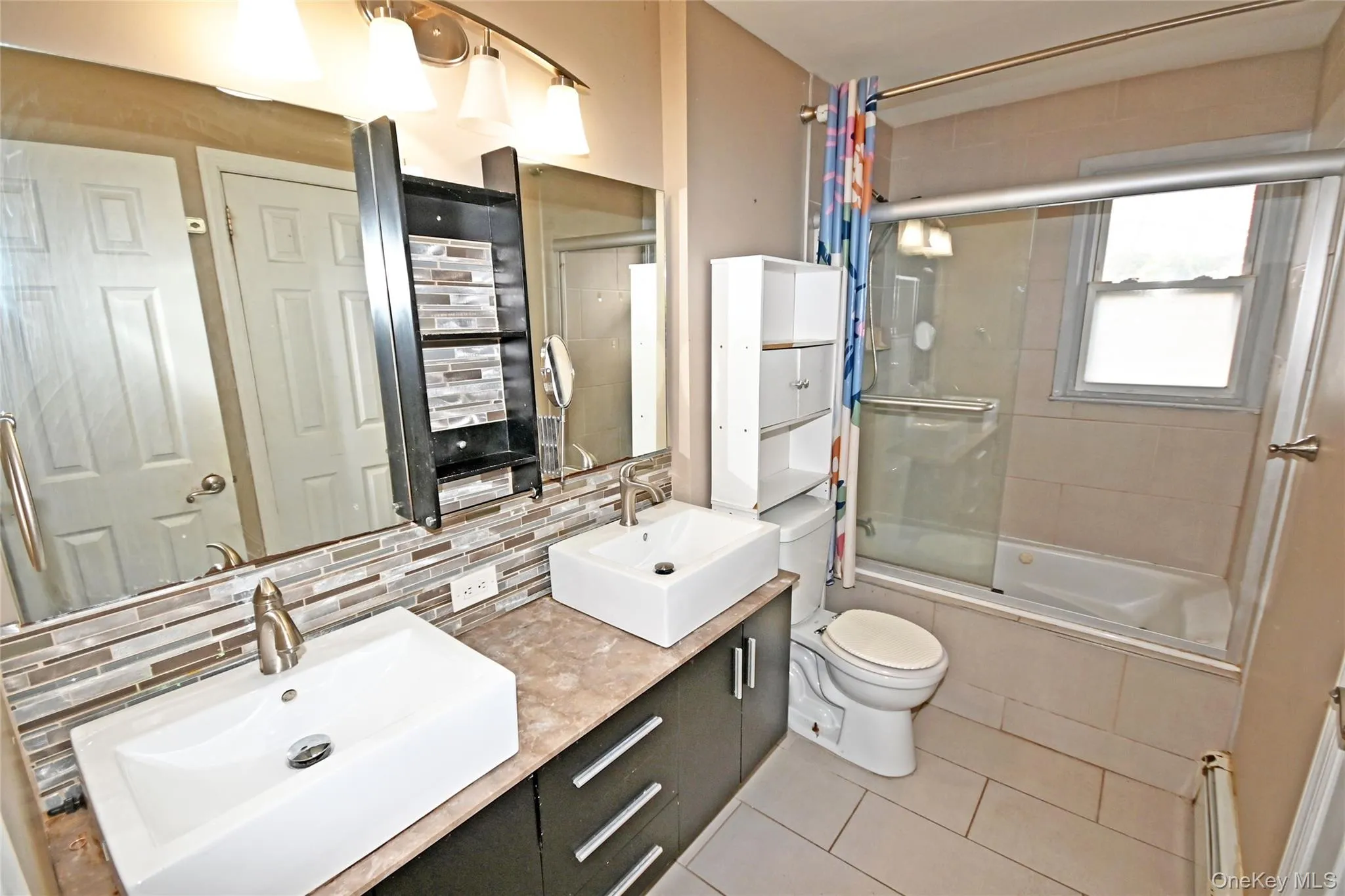 Full bath featuring double vanity, tiled shower / bath combo, decorative backsplash, light tile patterned floors, and a baseboard radiator Full bath featuring double vanity, tiled shower / bath combo, decorative backsplash, light tile patterned floors, and a baseboard radiator