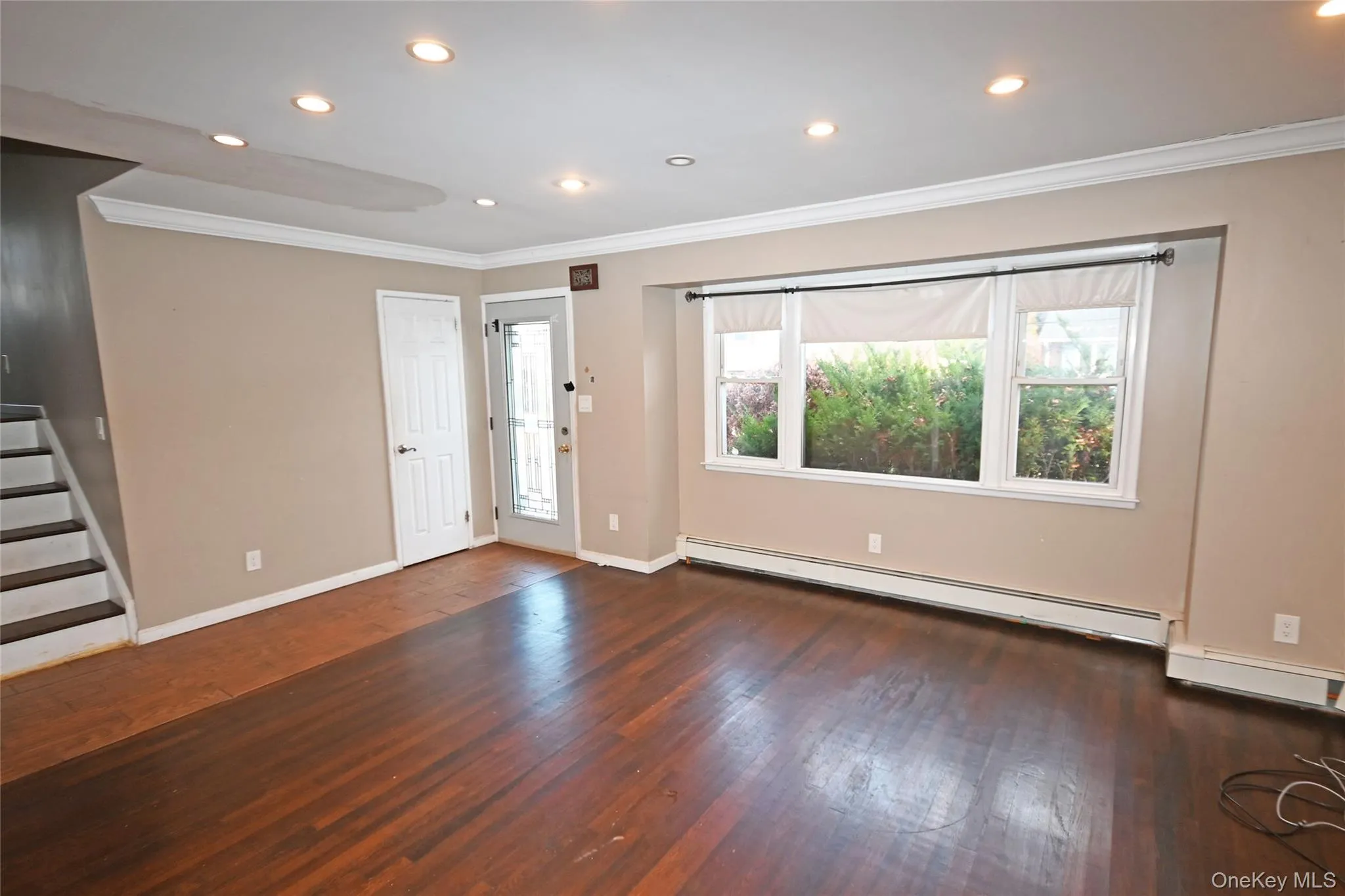 Unfurnished living room featuring crown molding, dark wood-type flooring, recessed lighting, a baseboard radiator, and stairway Unfurnished living room featuring crown molding, dark wood-type flooring, recessed lighting, a baseboard radiator, and stairway