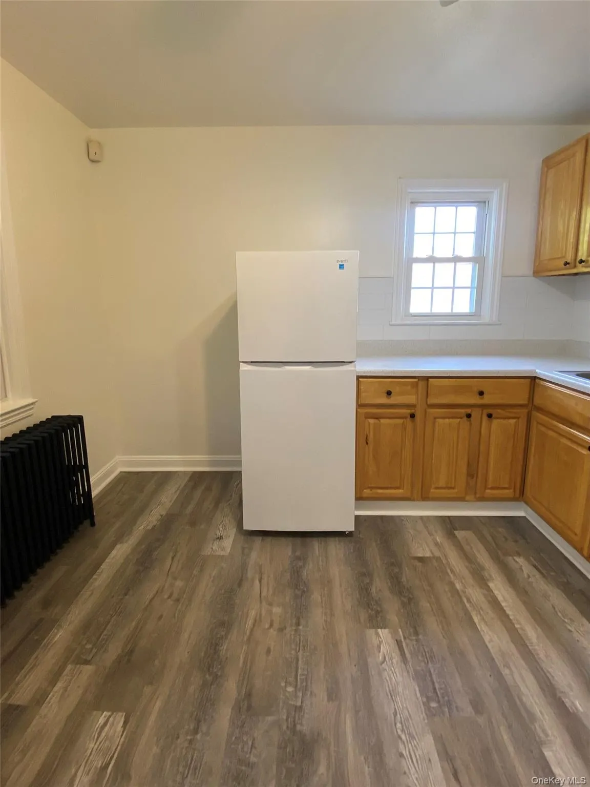 Kitchen featuring freestanding refrigerator, light countertops, radiator, dark wood-type flooring, and brown cabinets Kitchen featuring freestanding refrigerator, light countertops, radiator, dark wood-type flooring, and brown cabinets