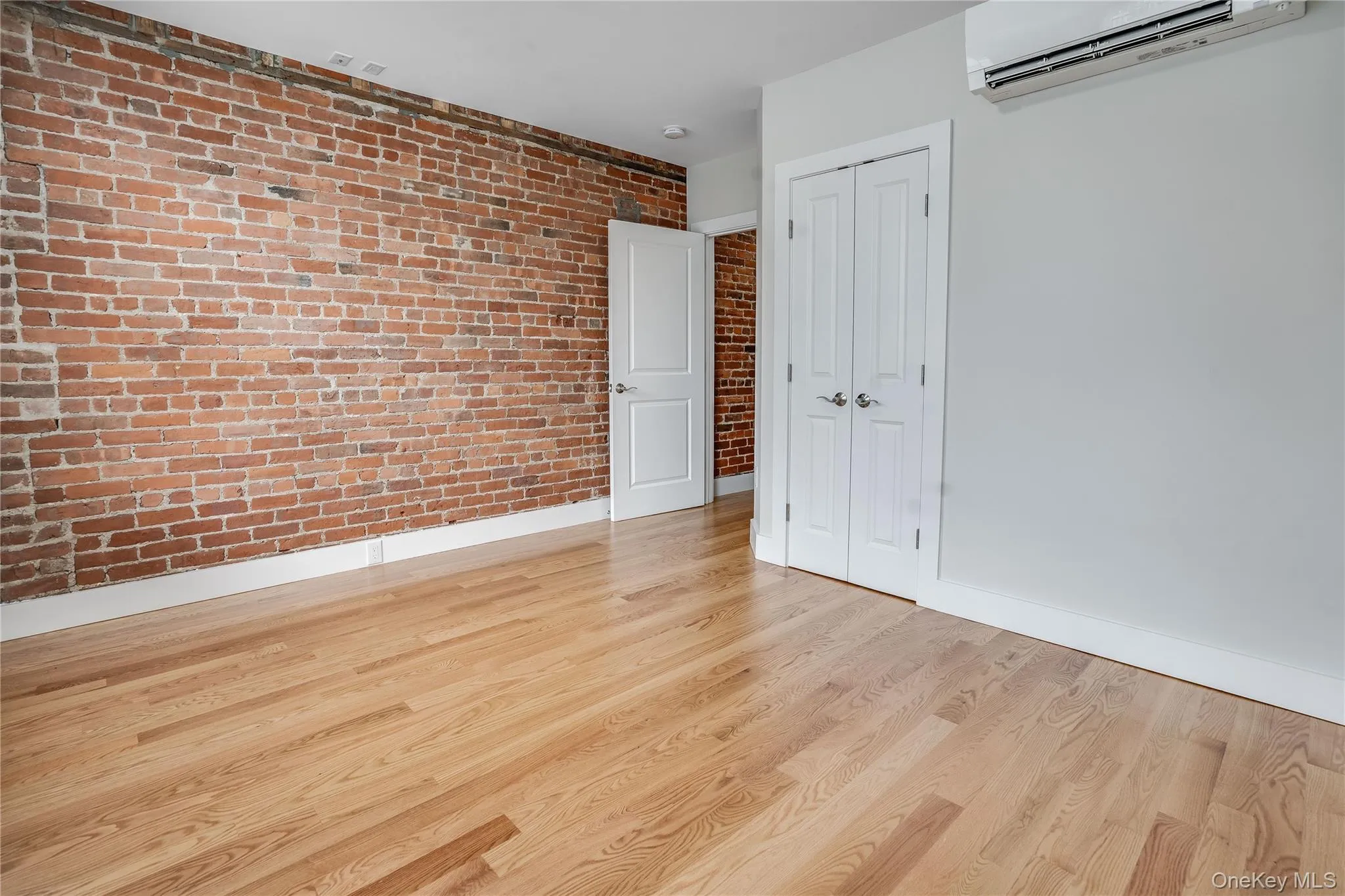 Unfurnished bedroom with a wall mounted air conditioner, light wood-type flooring, brick wall, and a closet Unfurnished bedroom with a wall mounted air conditioner, light wood-type flooring, brick wall, and a closet