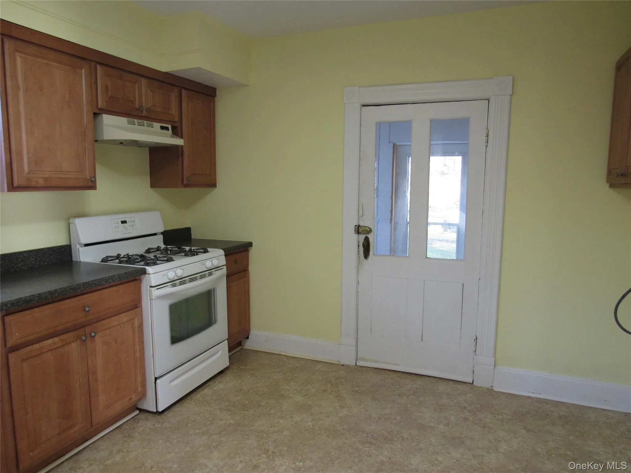 Kitchen with brown cabinetry, white gas stove, dark countertops, and under cabinet range hood Kitchen with brown cabinetry, white gas stove, dark countertops, and under cabinet range hood