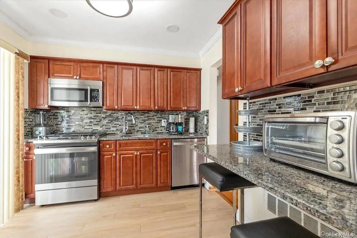 Kitchen with appliances with stainless steel finishes, dark stone countertops, tasteful backsplash, ornamental molding, and light wood-style flooring Kitchen with appliances with stainless steel finishes, dark stone countertops, tasteful backsplash, ornamental molding, and light wood-style flooring