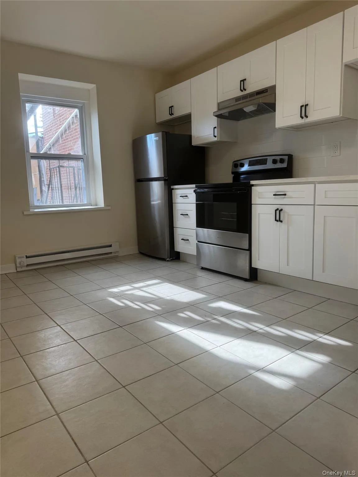 Kitchen with white cabinets, appliances with stainless steel finishes, light tile patterned floors, a baseboard radiator, and under cabinet range hood Kitchen with white cabinets, appliances with stainless steel finishes, light tile patterned floors, a baseboard radiator, and under cabinet range hood