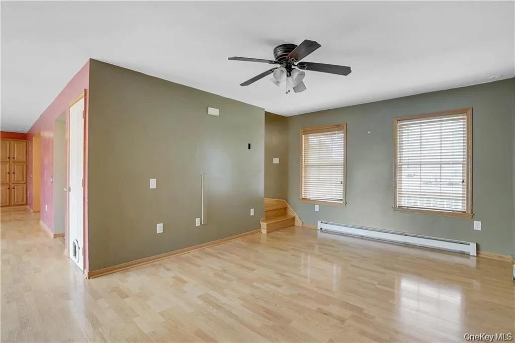 Spare room with baseboard heating, light wood finished floors, a ceiling fan, and stairs Spare room with baseboard heating, light wood finished floors, a ceiling fan, and stairs
