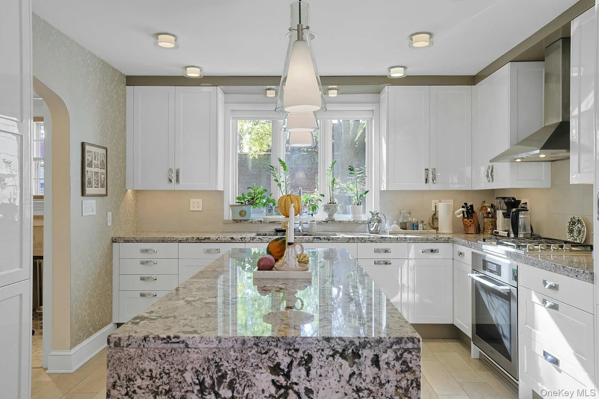 Kitchen with wall chimney range hood, light stone counters, hanging light fixtures, arched walkways, and white cabinetry Kitchen with wall chimney range hood, light stone counters, hanging light fixtures, arched walkways, and white cabinetry