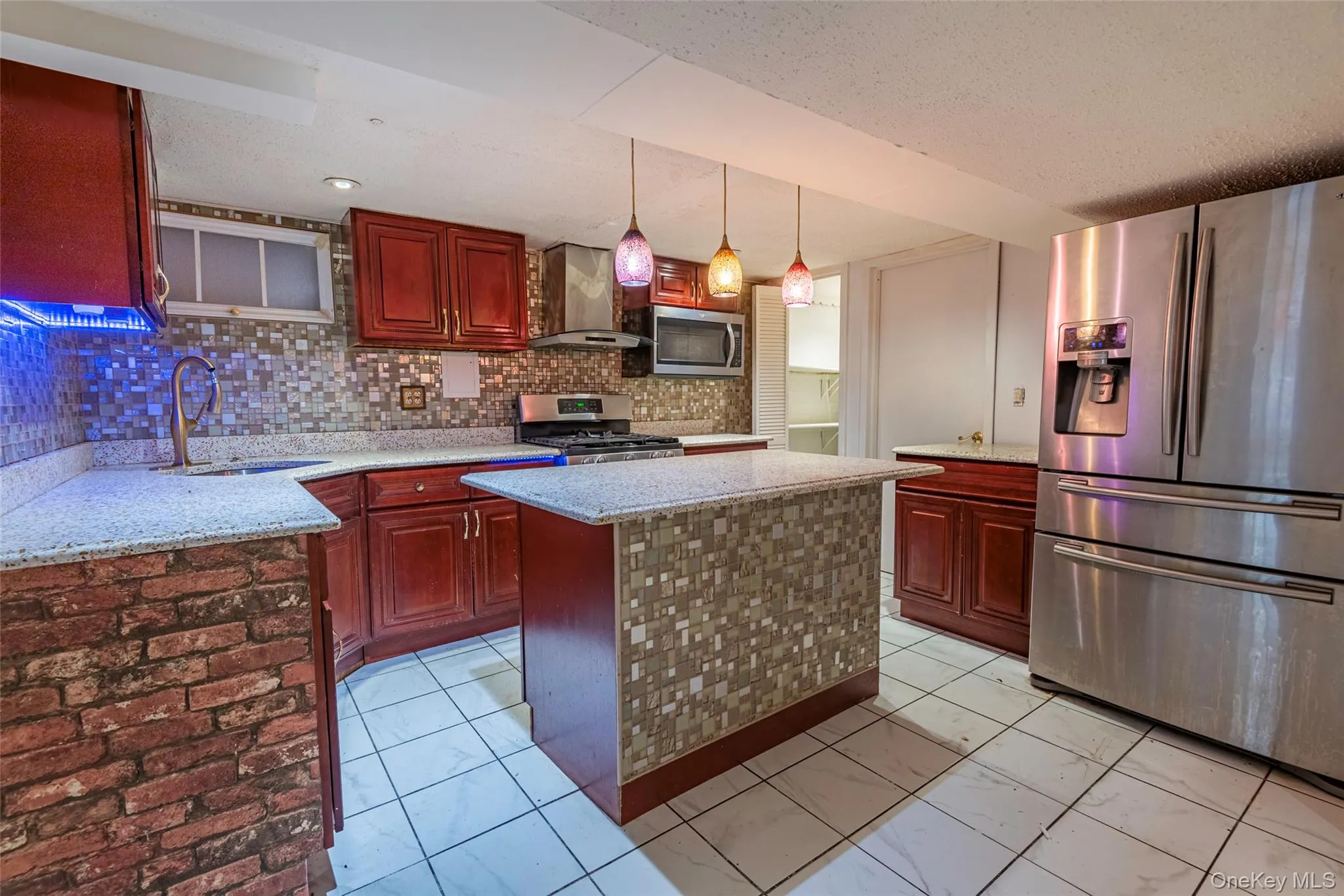 Kitchen featuring appliances with stainless steel finishes, decorative backsplash, wall chimney exhaust hood, hanging light fixtures, and light stone counters Kitchen featuring appliances with stainless steel finishes, decorative backsplash, wall chimney exhaust hood, hanging light fixtures, and light stone counters