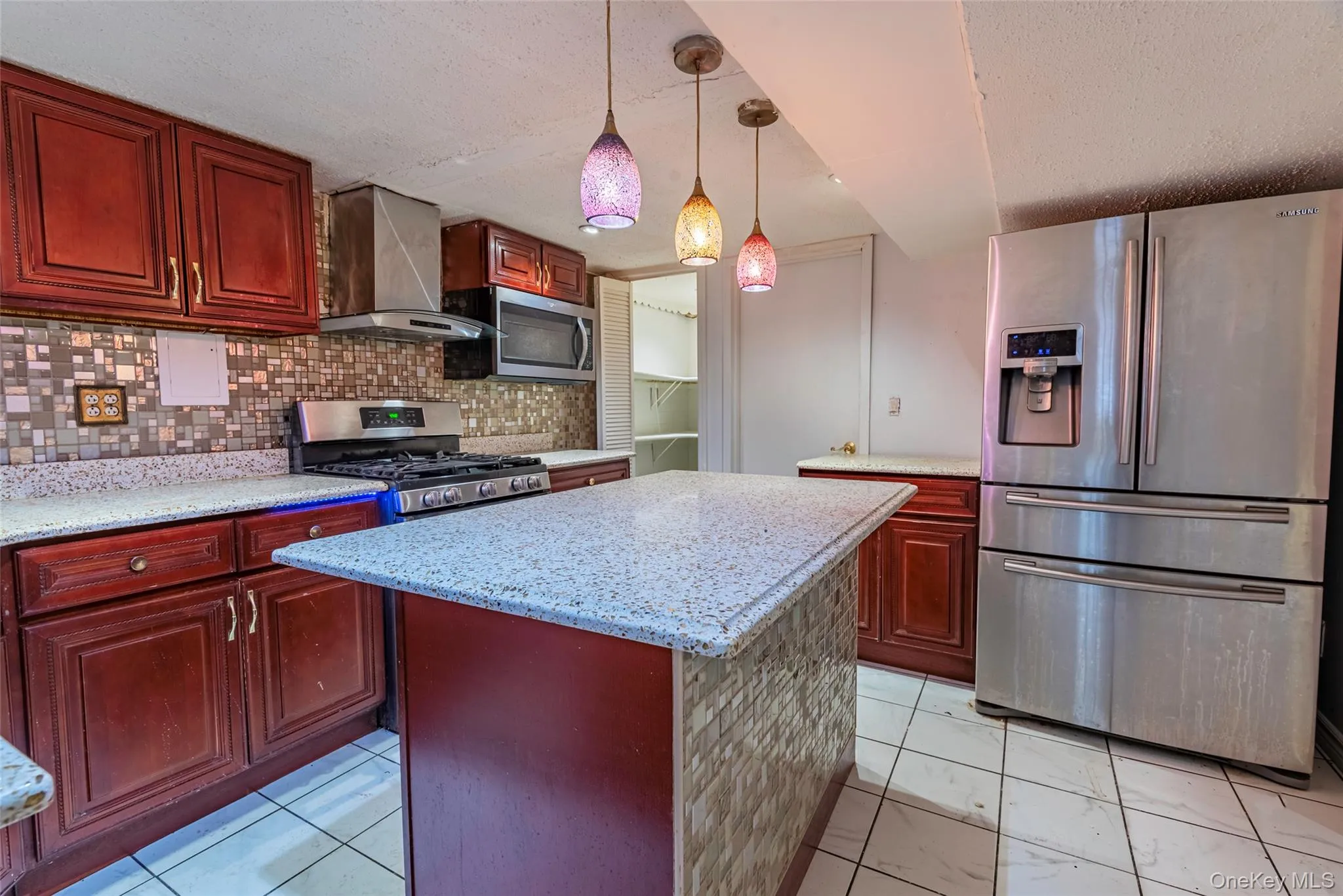 Kitchen with appliances with stainless steel finishes, hanging light fixtures, backsplash, ventilation hood, and a textured ceiling Kitchen with appliances with stainless steel finishes, hanging light fixtures, backsplash, ventilation hood, and a textured ceiling