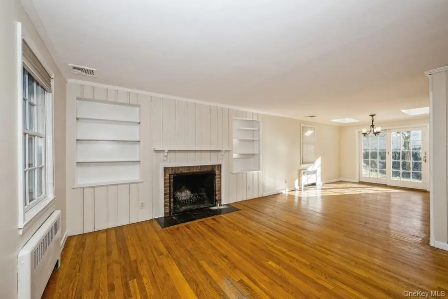 Unfurnished living room featuring radiator, light wood finished floors, built in shelves, crown molding, and a chandelier Unfurnished living room featuring radiator, light wood finished floors, built in shelves, crown molding, and a chandelier