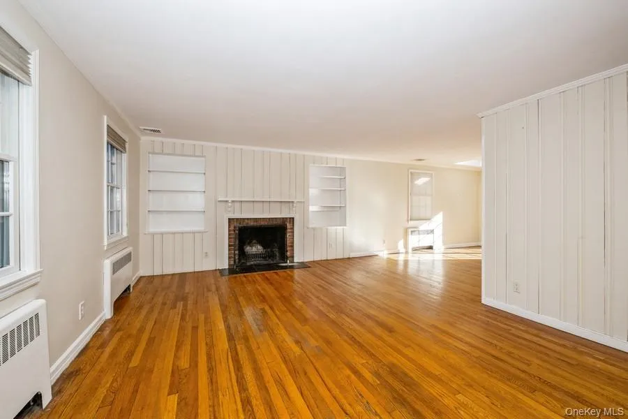 Unfurnished living room with radiator heating unit, crown molding, wood finished floors, a brick fireplace, and built in features Unfurnished living room with radiator heating unit, crown molding, wood finished floors, a brick fireplace, and built in features