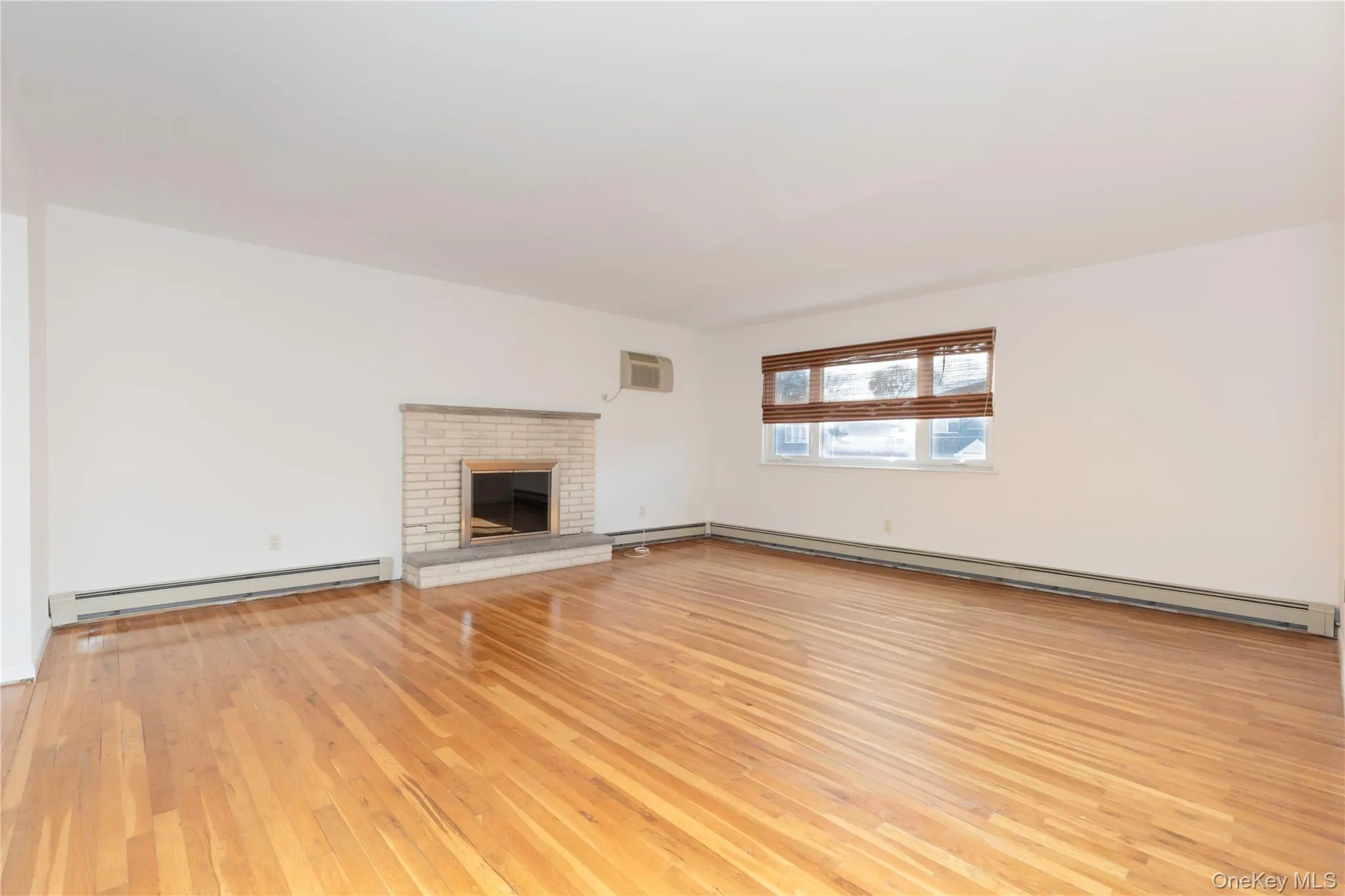 living room featuring a baseboard radiator, light wood-style flooring, a brick fireplace, and a wall mounted AC living room featuring a baseboard radiator, light wood-style flooring, a brick fireplace, and a wall mounted AC