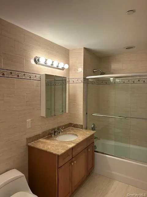 Full bath featuring tile walls, enclosed tub / shower combo, vanity, and light tile patterned floors Full bath featuring tile walls, enclosed tub / shower combo, vanity, and light tile patterned floors
