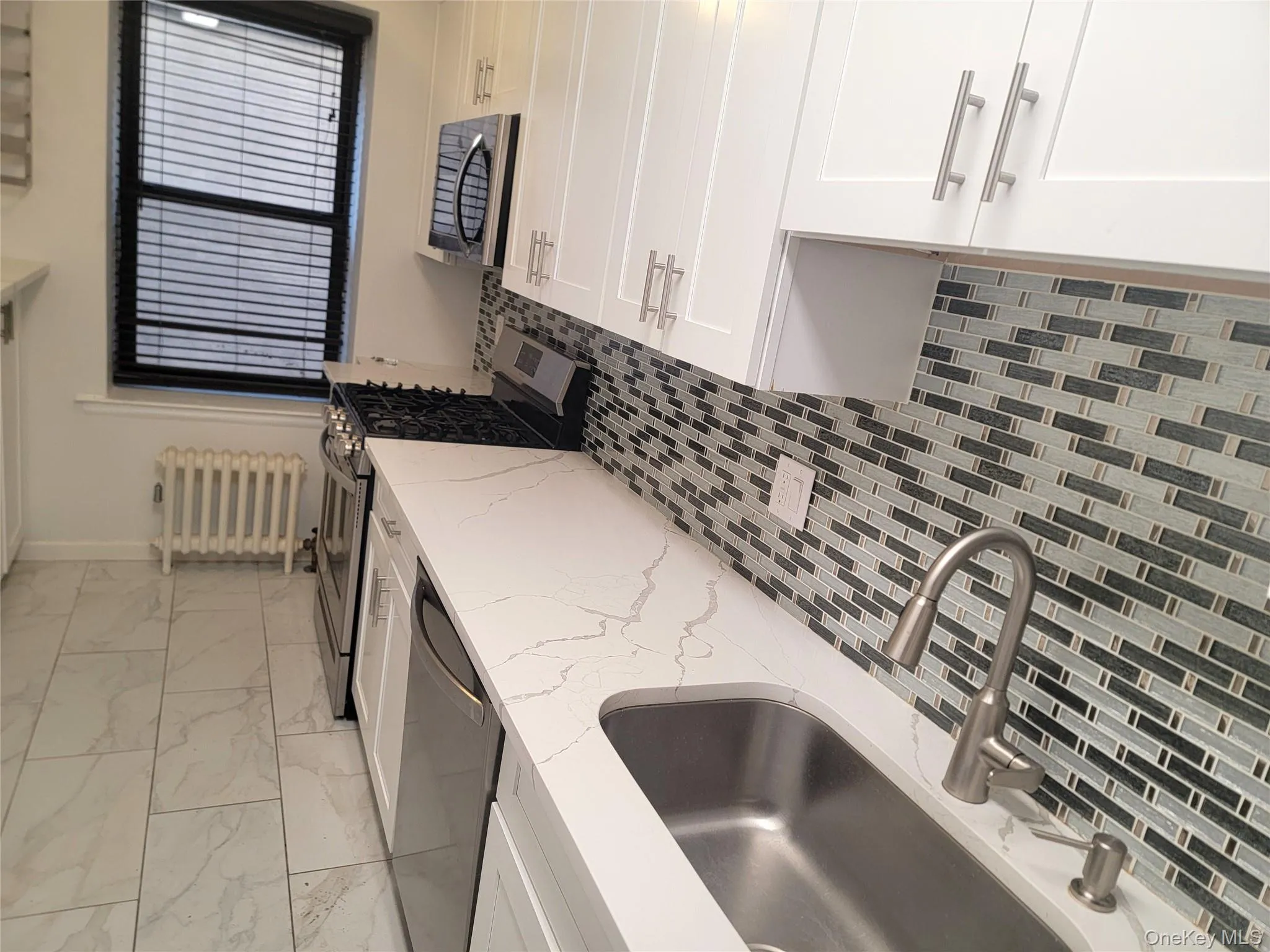 Kitchen with backsplash, stainless steel appliances, radiator, light stone countertops, and white cabinetry Kitchen with backsplash, stainless steel appliances, radiator, light stone countertops, and white cabinetry