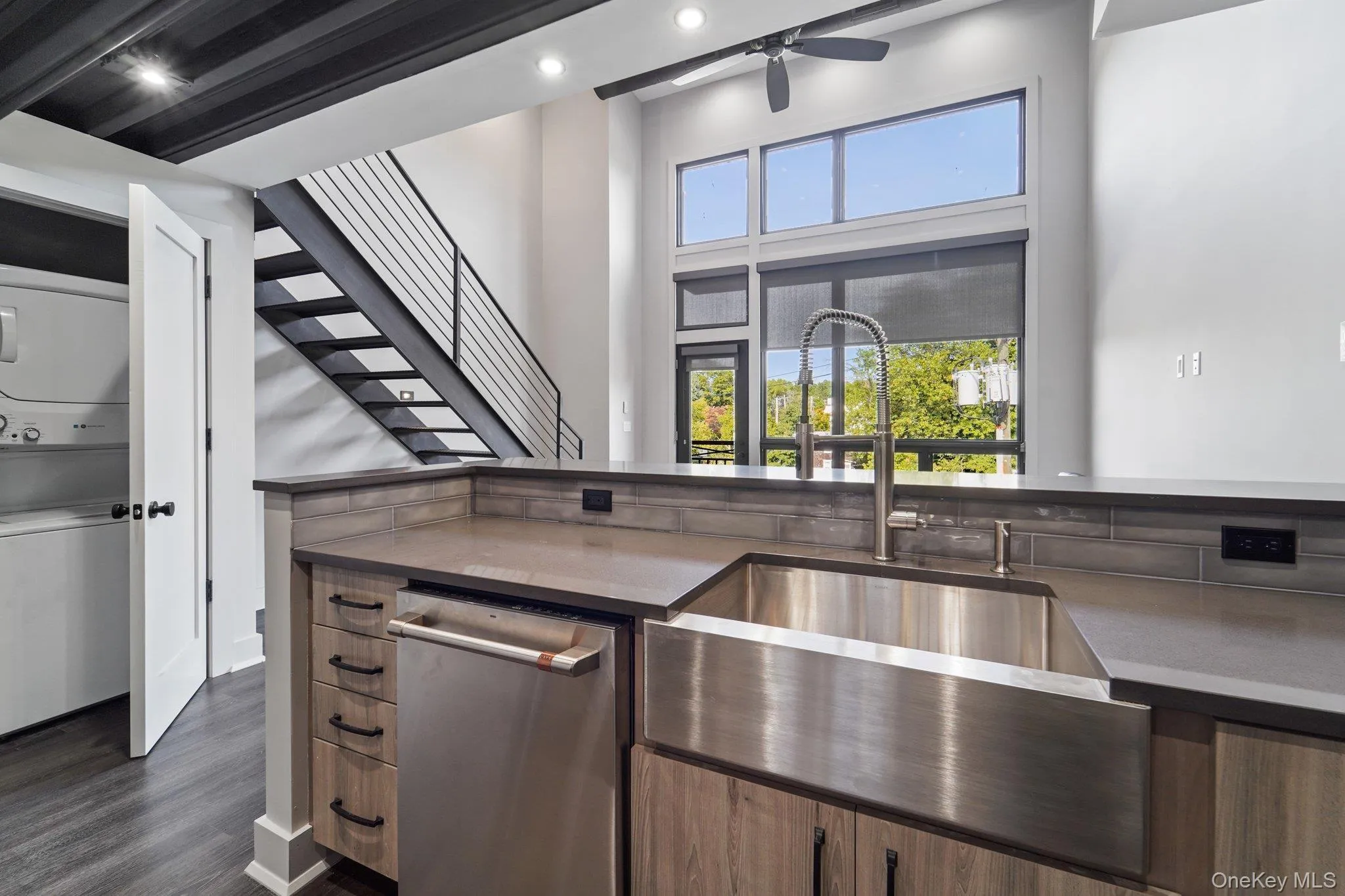 Kitchen with stainless steel dishwasher, stacked washer and clothes dryer, dark wood-style flooring, a ceiling fan, and dark stone countertops Kitchen with stainless steel dishwasher, stacked washer and clothes dryer, dark wood-style flooring, a ceiling fan, and dark stone countertops