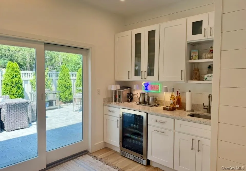 Indoor wet bar featuring white cabinets, wine cooler, light wood-style floors, light stone counters, and glass insert cabinets Indoor wet bar featuring white cabinets, wine cooler, light wood-style floors, light stone counters, and glass insert cabinets