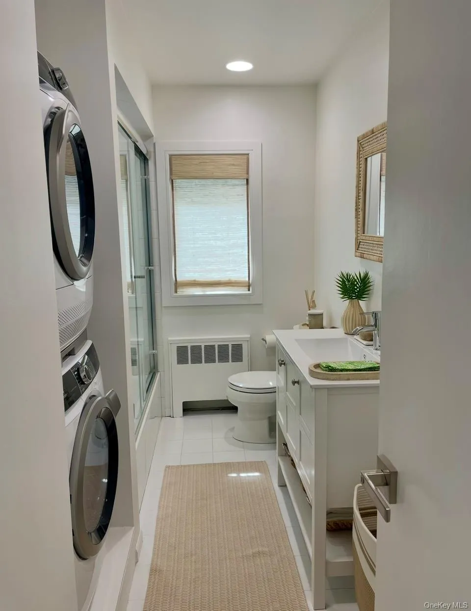 Full bath with vanity, radiator heating unit, stacked washer and clothes dryer, light tile patterned floors, and bath / shower combo with glass door Full bath with vanity, radiator heating unit, stacked washer and clothes dryer, light tile patterned floors, and bath / shower combo with glass door