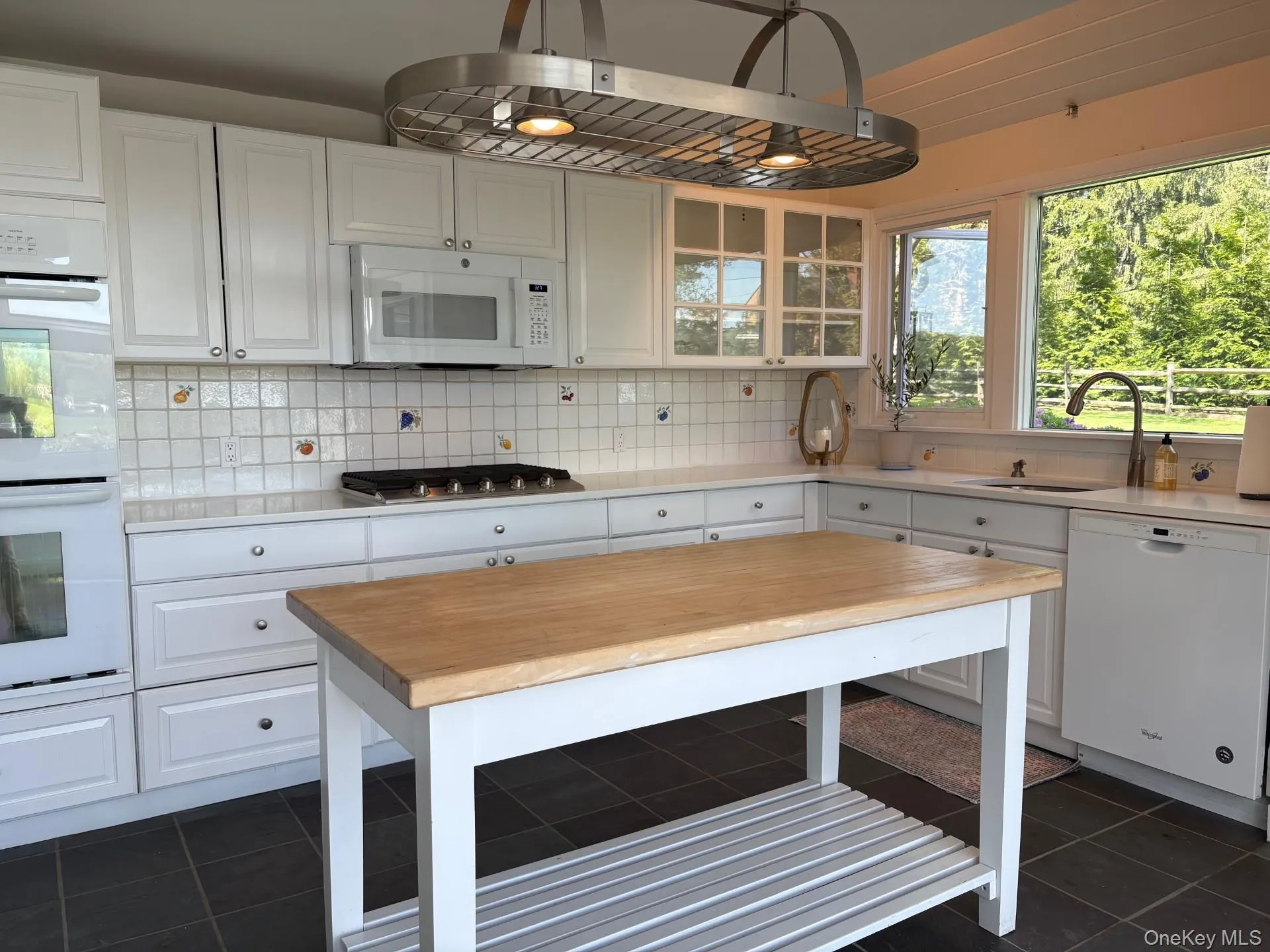 Kitchen featuring white cabinetry, a central island with a wood-finish countertop, integrated stainless steel appliances, a white tile backsplash, and dark tile flooring Kitchen featuring white cabinetry, a central island with a wood-finish countertop, integrated stainless steel appliances, a white tile backsplash, and dark tile flooring