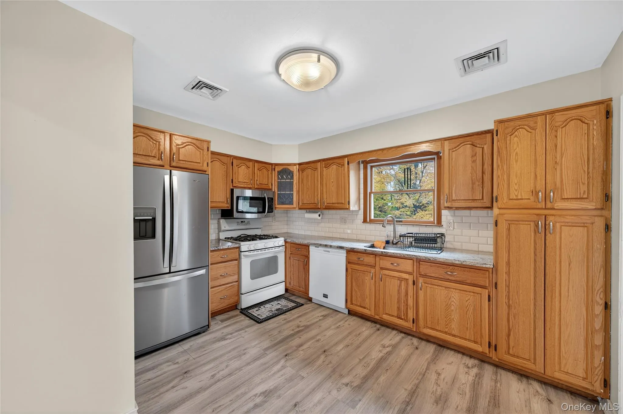 Kitchen with appliances with stainless steel finishes, light wood-style floors, decorative backsplash, glass insert cabinets, and brown cabinets Kitchen with appliances with stainless steel finishes, light wood-style floors, decorative backsplash, glass insert cabinets, and brown cabinets