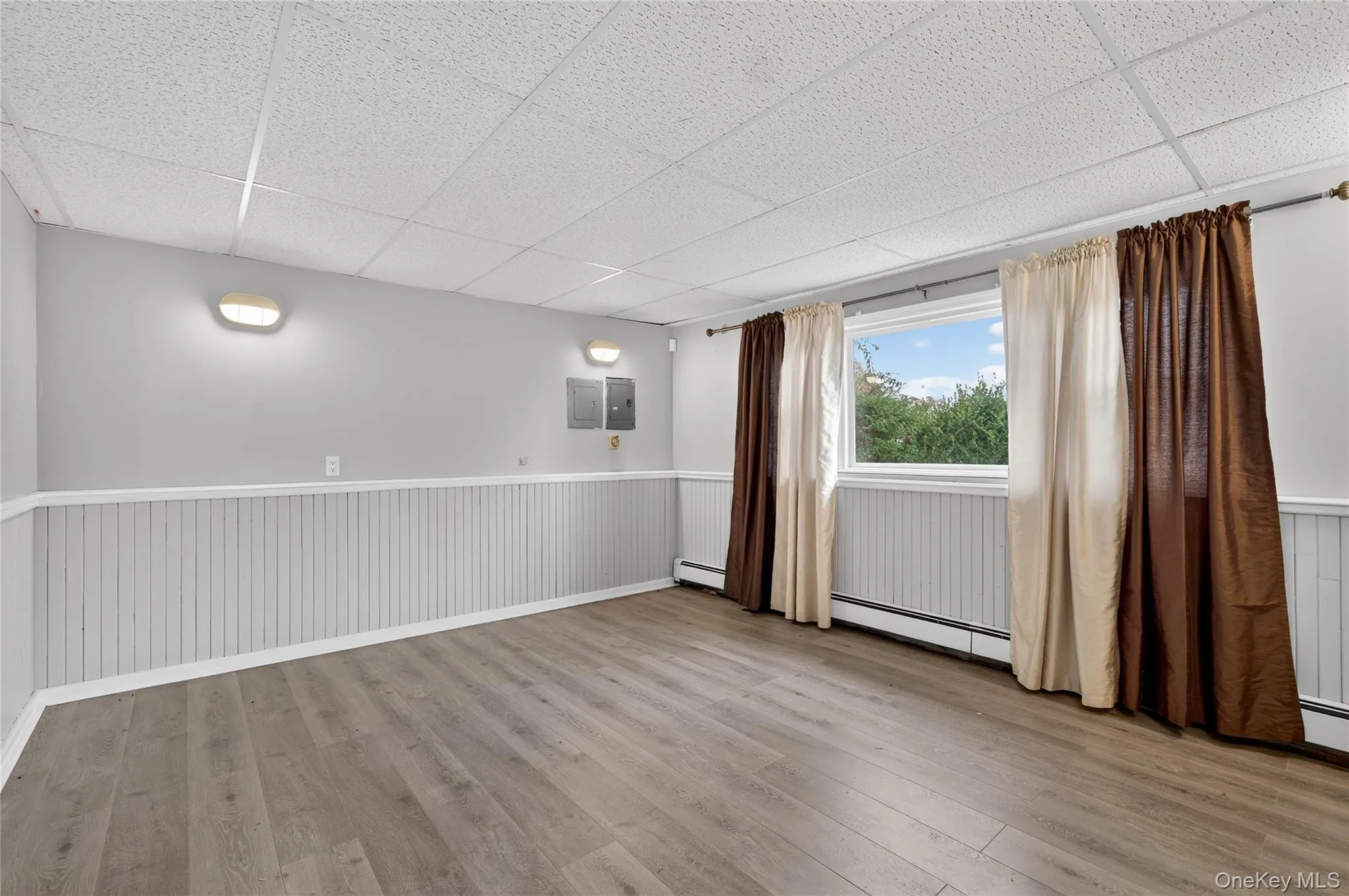 Spare room with light wood finished floors, a paneled ceiling, a baseboard heating unit, wainscoting, and electric panel Spare room with light wood finished floors, a paneled ceiling, a baseboard heating unit, wainscoting, and electric panel