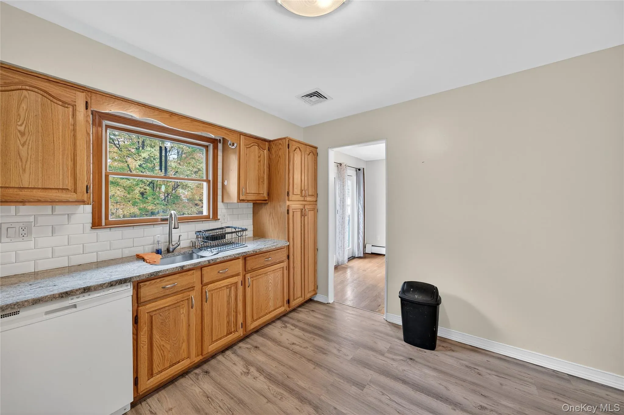 Kitchen with white dishwasher, backsplash, light wood-type flooring, brown cabinetry, and light stone counters Kitchen with white dishwasher, backsplash, light wood-type flooring, brown cabinetry, and light stone counters
