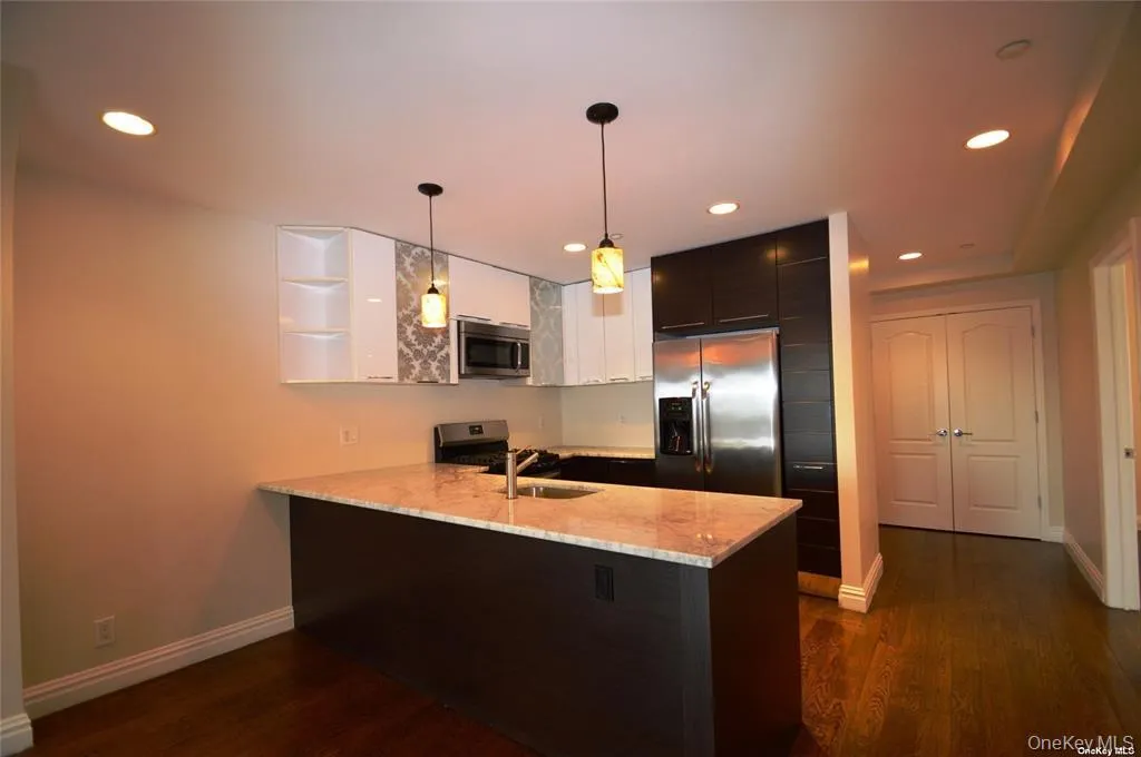Kitchen with light stone counters, appliances with stainless steel finishes, a peninsula, hanging light fixtures, and dark wood finished floors Kitchen with light stone counters, appliances with stainless steel finishes, a peninsula, hanging light fixtures, and dark wood finished floors