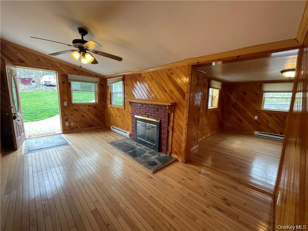 Unfurnished living room featuring wooden walls, hardwood / wood-style floors, a fireplace, ceiling fan, and baseboard heating Unfurnished living room featuring wooden walls, hardwood / wood-style floors, a fireplace, ceiling fan, and baseboard heating