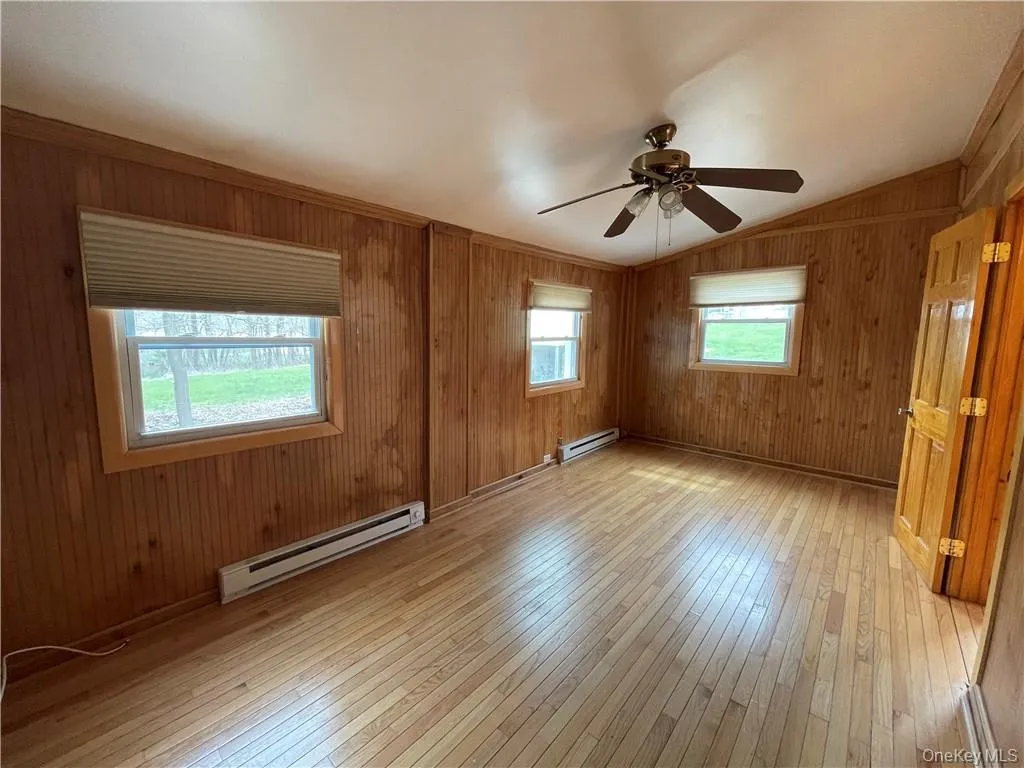 Spare room with a baseboard heating unit, wood walls, light wood-type flooring, vaulted ceiling, and a ceiling fan Spare room with a baseboard heating unit, wood walls, light wood-type flooring, vaulted ceiling, and a ceiling fan