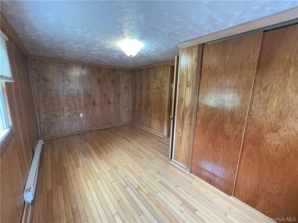 Unfurnished bedroom featuring light wood-type flooring, a baseboard heating unit, wood walls, a closet, and ornamental molding Unfurnished bedroom featuring light wood-type flooring, a baseboard heating unit, wood walls, a closet, and ornamental molding